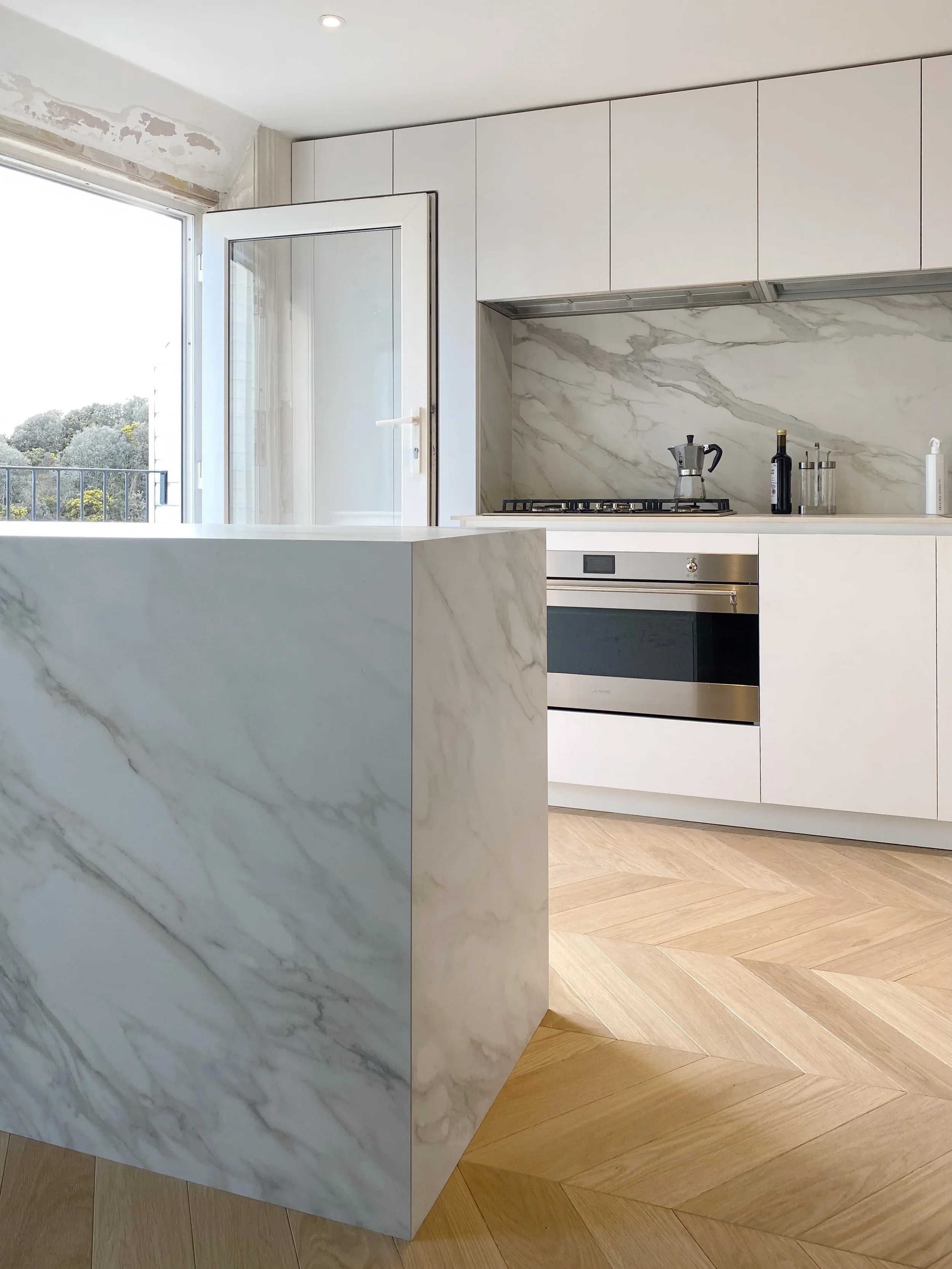 Contemporary bright kitchen featuring a monolithic central island and a matching rear splashback custom fabricated from white Calacatta marble-effect porcelain slabs, integrated with sleek handleless white cabinetry and light oak herringbone flooring