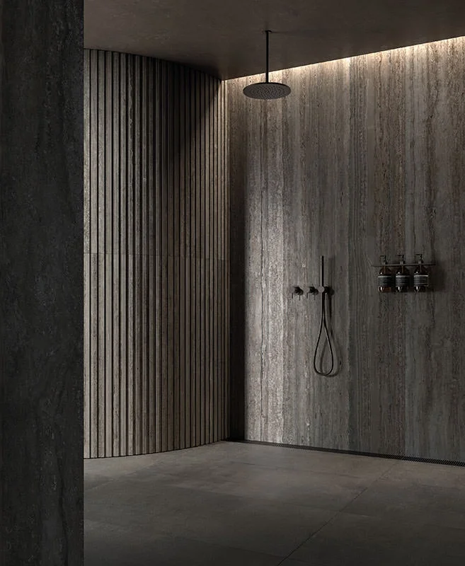 A modern shower featuring the Nativa collection from Kronos Ceramiche, utilizing large-format travertine-style porcelain alongside a curved fluted wall feature and matte black fixtures, available through PietraCasa.