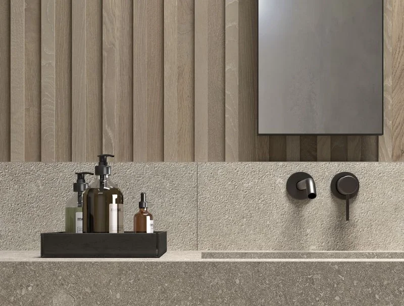 A bathroom vanity featuring Kronos Ceramiche's 'Le Reverse' porcelain stoneware in Dune, showcasing seamless large-format surfaces, an integrated washbasin, a matte black faucet, and natural oak-slat wall paneling, available through PietraCasa.