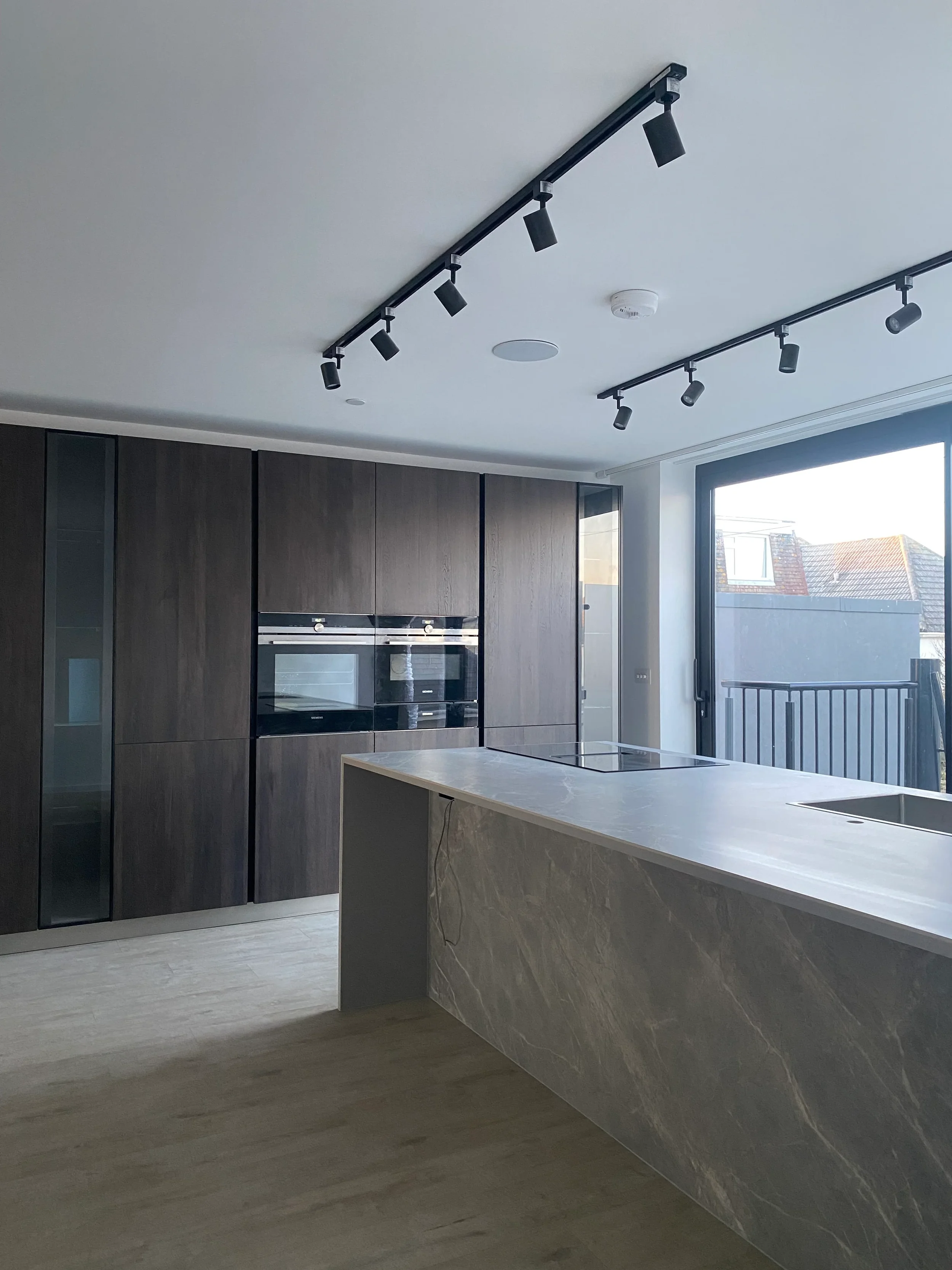Modern kitchen installation featuring a large central island with waterfall edges custom fabricated from light grey marble-effect porcelain slabs, integrated with a flush hob and undermount sink, set against dark wood floor-to-ceiling cabinets and bl