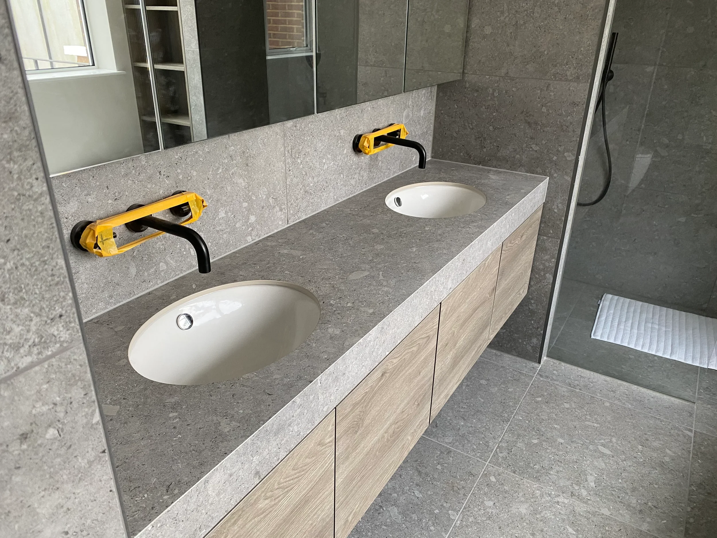 A high-end double vanity installation featuring seamless, large-format grey porcelain countertops and backsplash, paired with wood-finish cabinetry and matte black hardware.