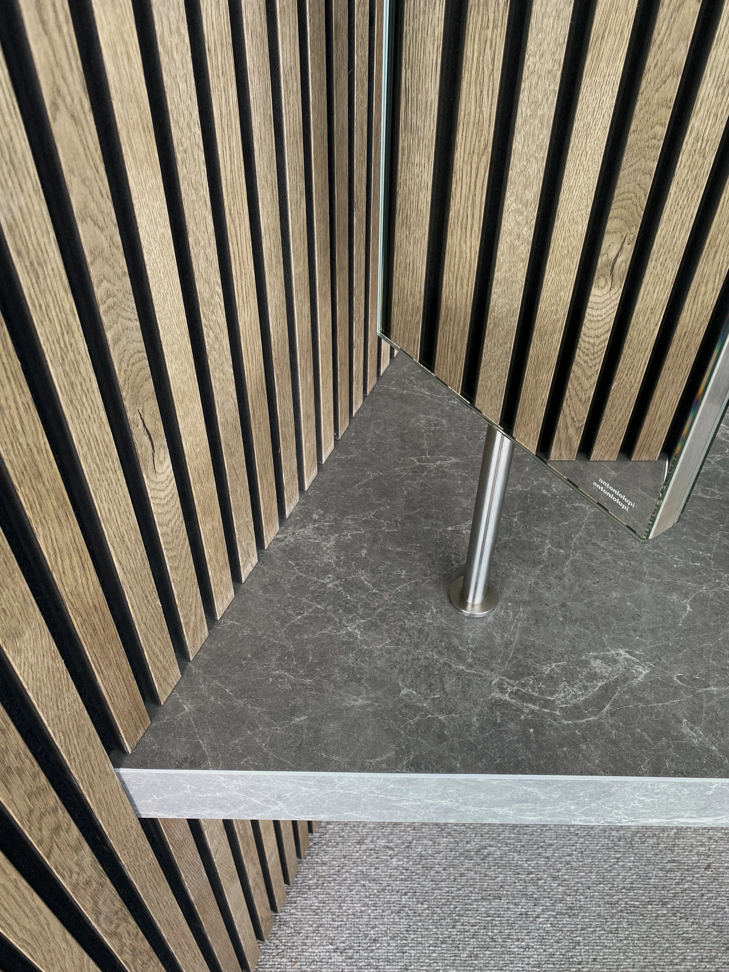 Close-up of a luxury antoniolupi mirror mounted on a dark grey marble-effect porcelain worktop by PietraCasa, framed by contemporary oak slat wall panelling.