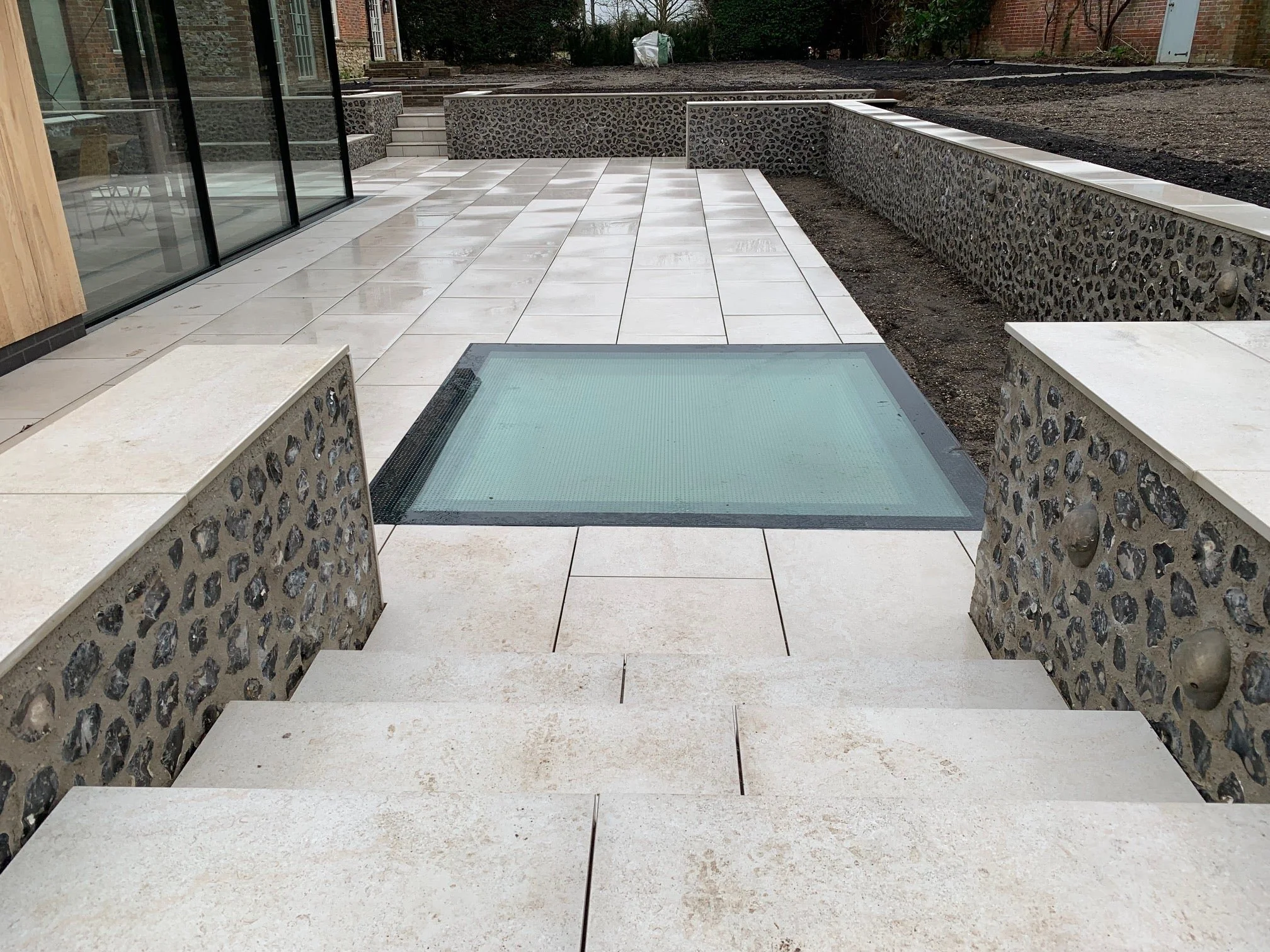 Luxury terrace featuring light-colored porcelain paving tiles integrated with a flush-mounted glass floor lightwell and traditional flint masonry walls.