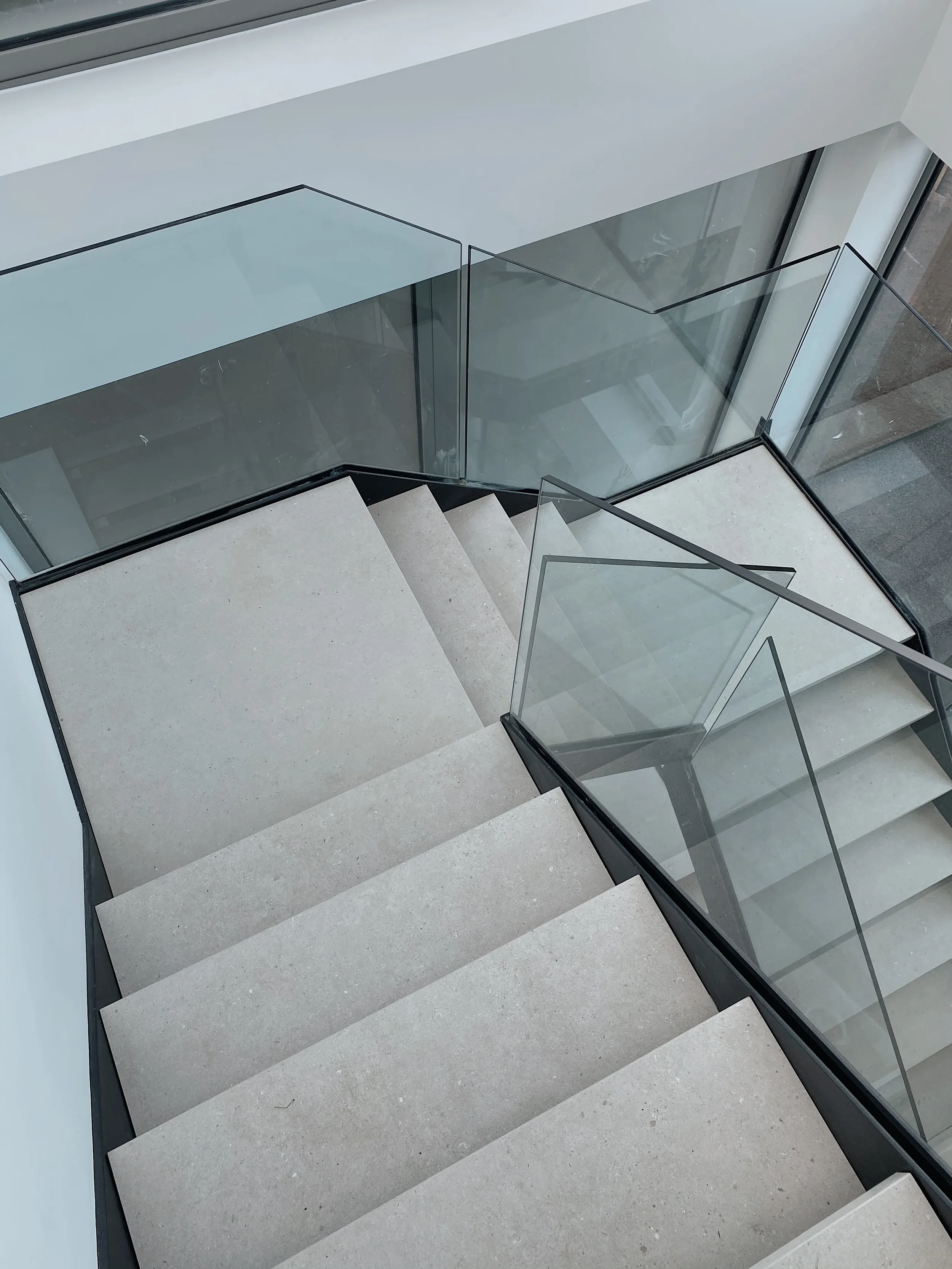 A luxury contemporary multi-flight staircase featuring custom-fabricated steps and half-landings clad in matte cream stone-effect porcelain slabs, paired with a modern black-framed glass balustrade.