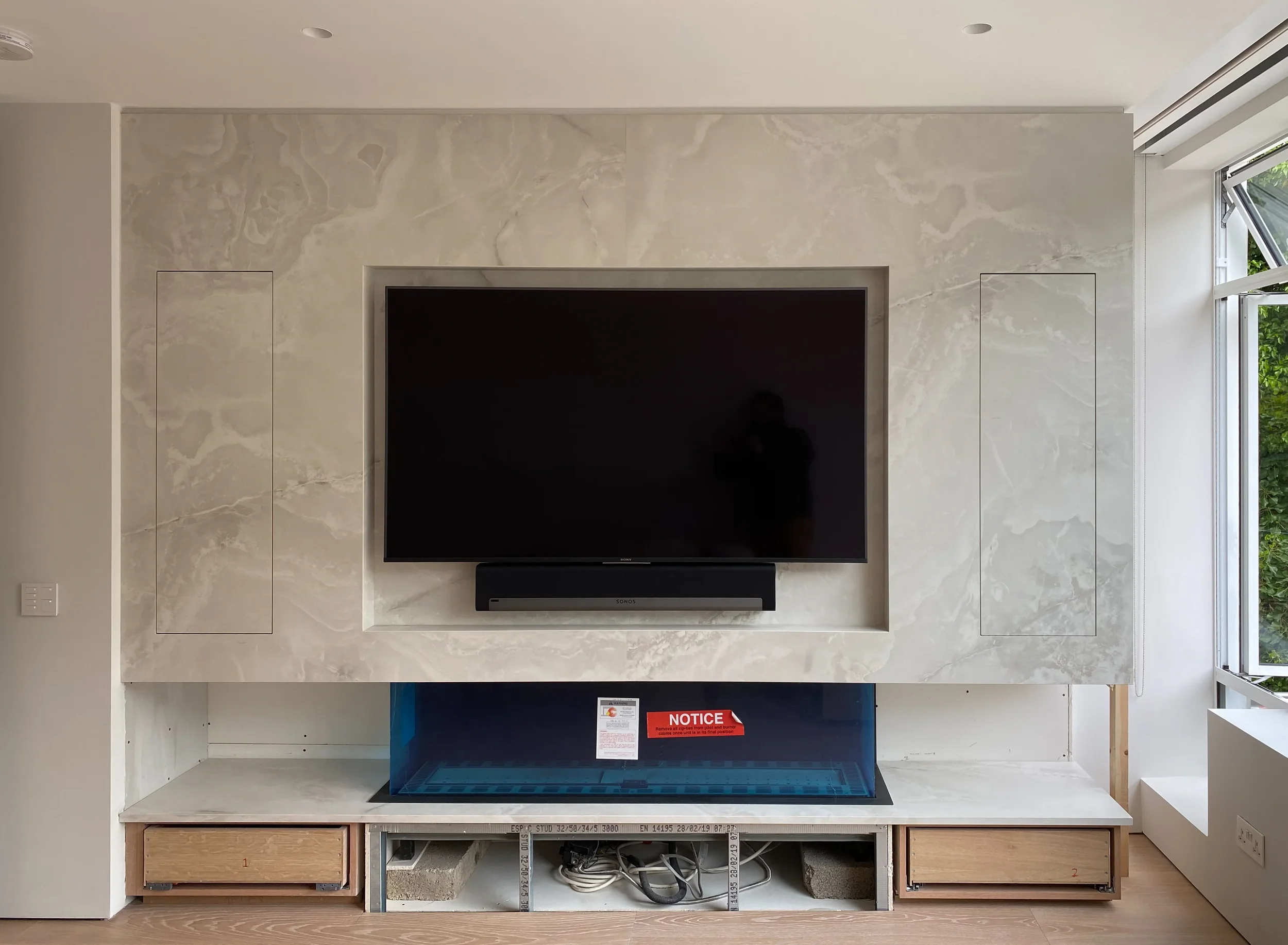 High-end minimalist living room feature wall under construction, showcasing a bookmatched translucent stone-look porcelain cladding system over an integrated modern linear fireplace and low-profile media bench.