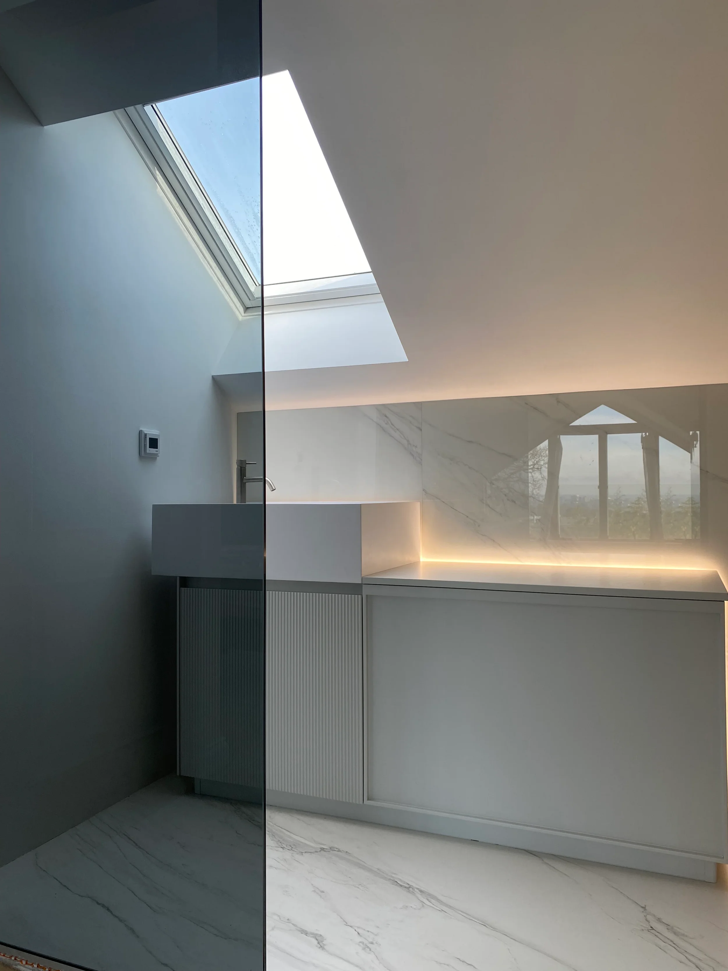 White fluted bathroom vanity and low cabinets with an LED-illuminated marble-effect porcelain panel under a loft skylight
