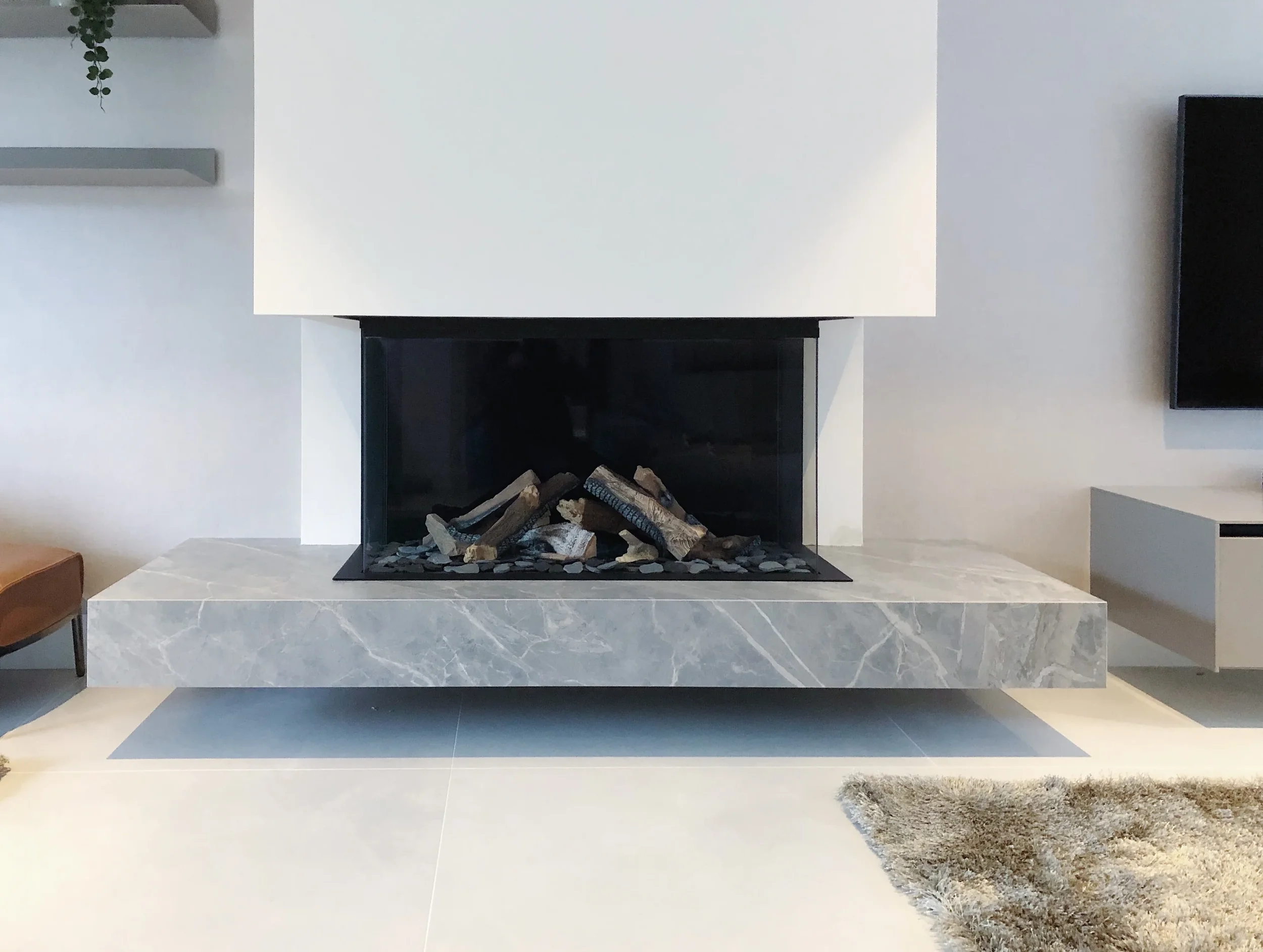 A luxury contemporary living room feature wall showcasing a low-profile cantilevered floating hearth custom-fabricated from grey marble-effect porcelain slabs, integrated with a modern three-sided glass gas fireplace.