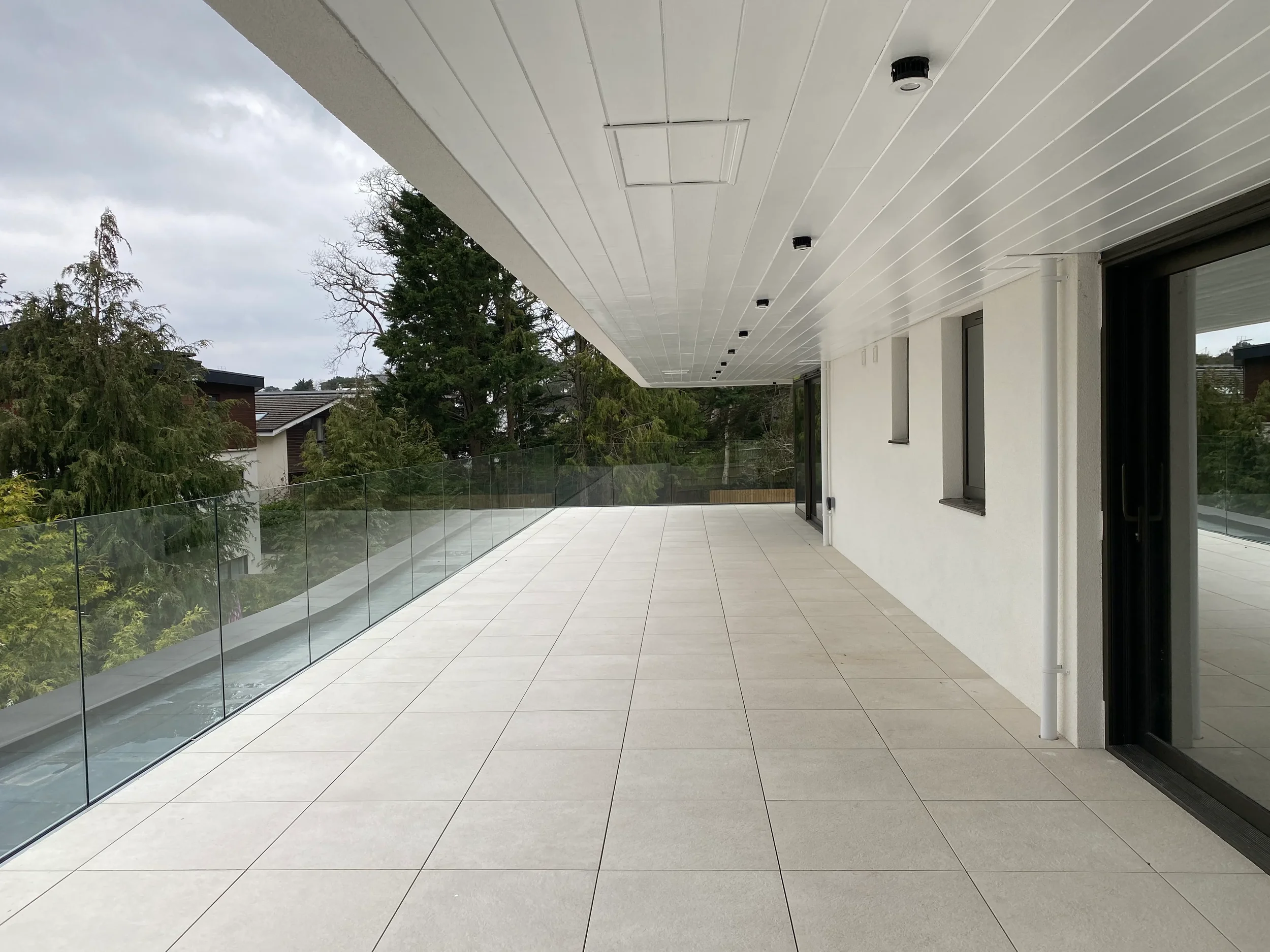 Wide-angle view of a luxury balcony featuring light-colored porcelain floor tiles, a frameless glass balustrade, and a modern white soffit ceiling.
