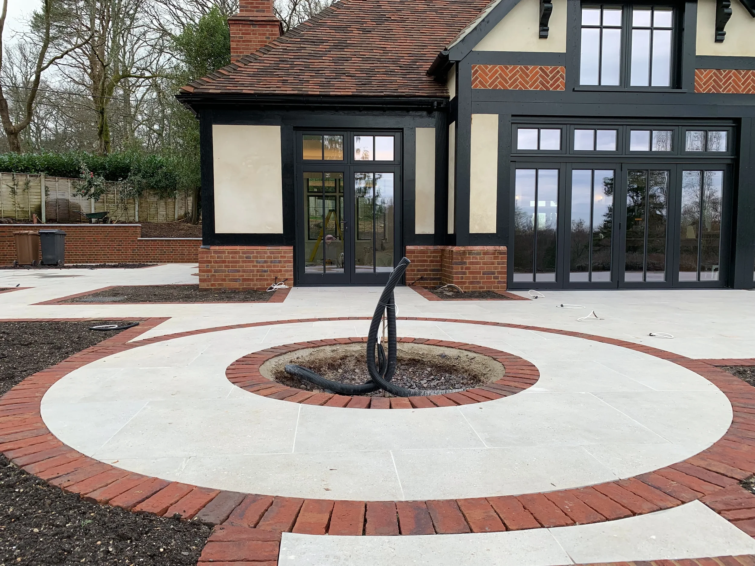 Circular porcelain patio feature bordered by heritage red bricks, situated in a garden setting outside a traditional mock-Tudor home.