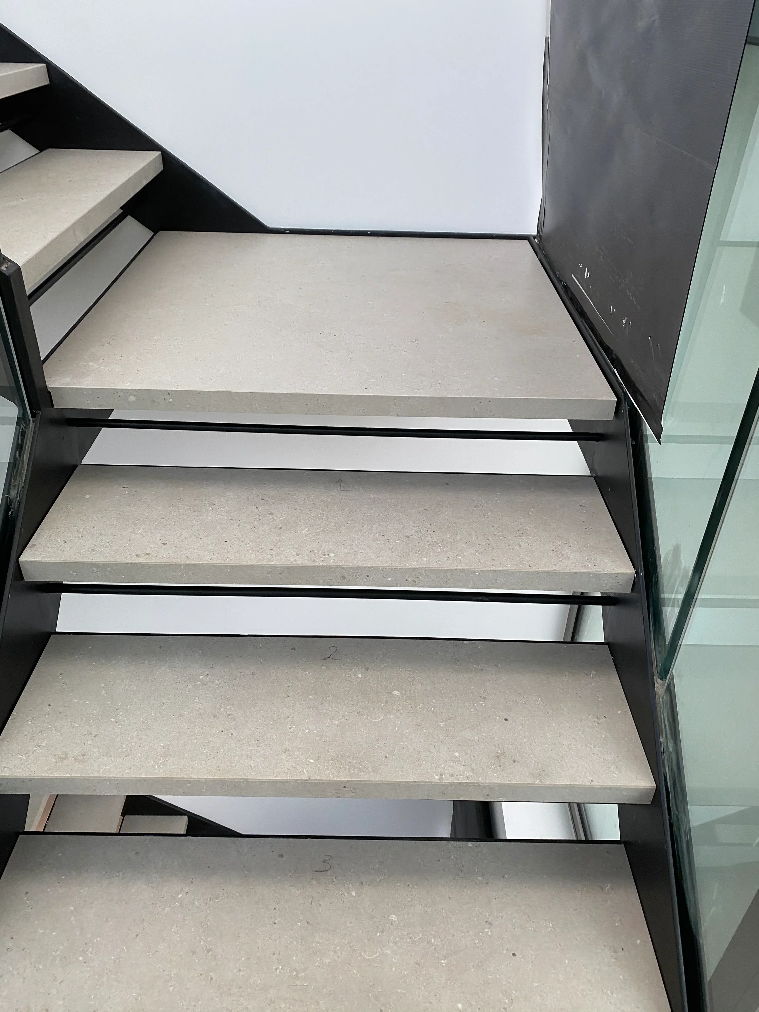 Straight-on view of an open-tread staircase featuring custom-fabricated matte cream porcelain steps and a half-landing set into a black steel frame.