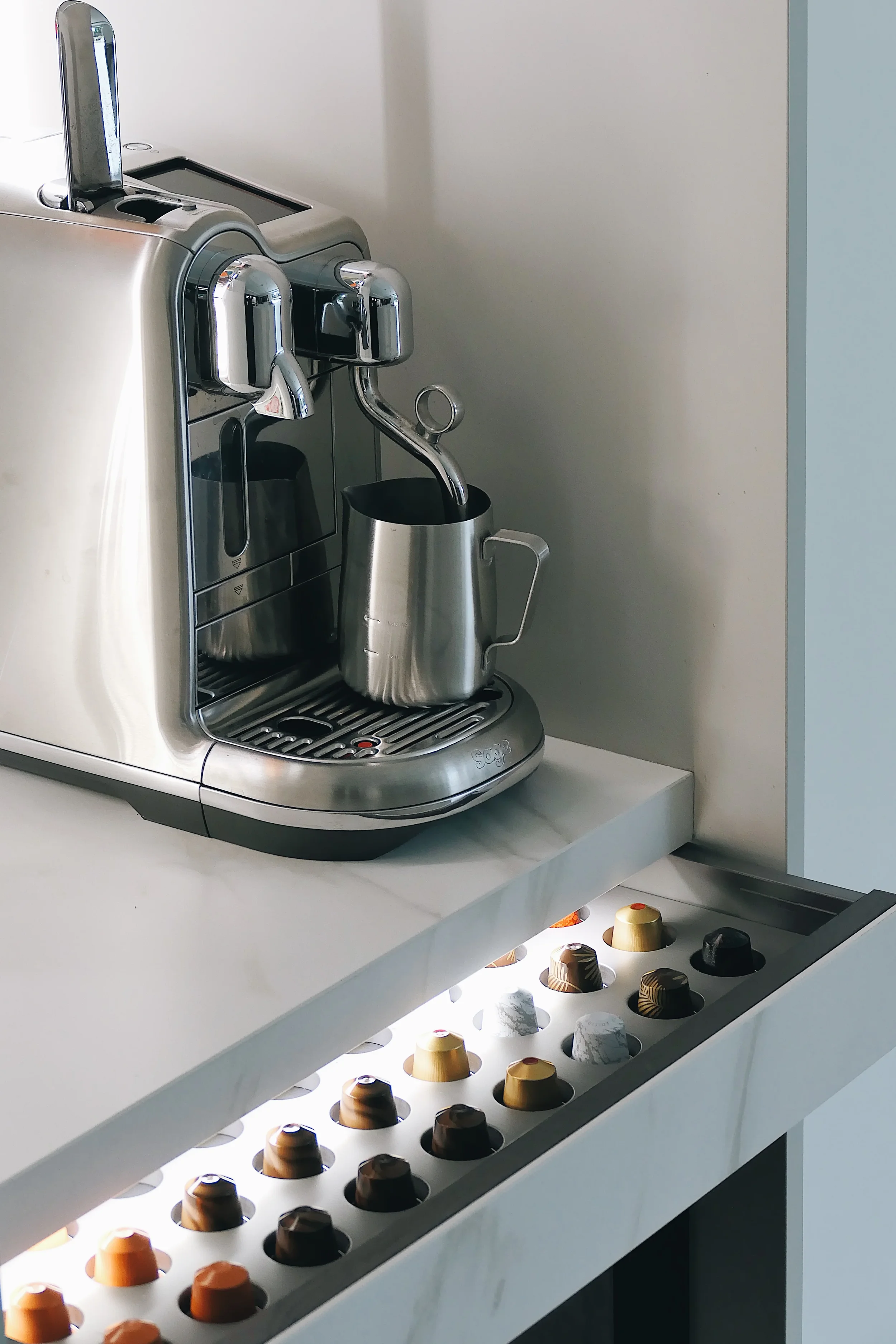 Luxury coffee station featuring a stainless steel Sage espresso machine on a white marble-effect porcelain slab worktop, highlighting a custom integrated pull-out drawer organizer with built-in LED lighting for coffee capsules.