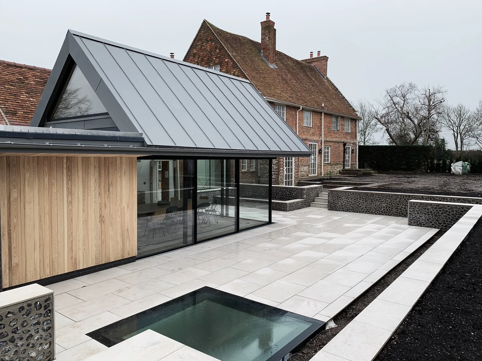 Architectural juxtaposition of a sleek, modern glass-and-metal home addition against a traditional brick residence, complemented by light-colored porcelain paving