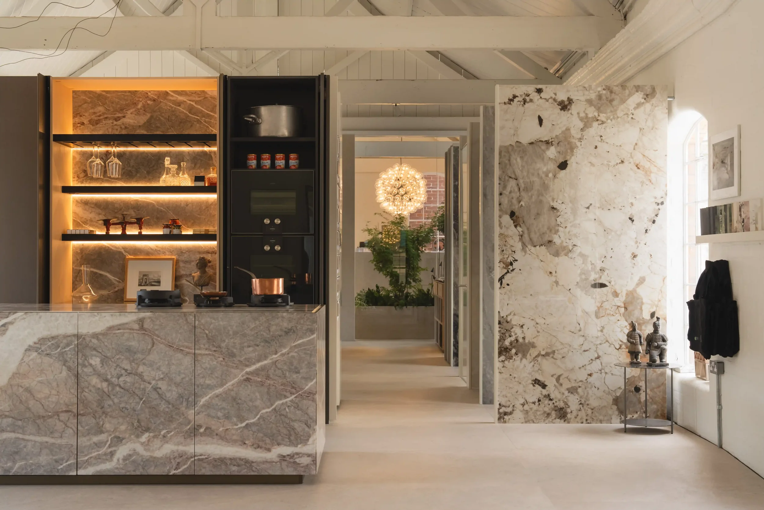 Interior view of the shared Abitalia and PietraCasa showroom, showcasing a luxury designer kitchen island and illuminated pocket-door larder clad in heavily veined grey marble-look porcelain slabs alongside a large-format Patagonia-effect porcelain f