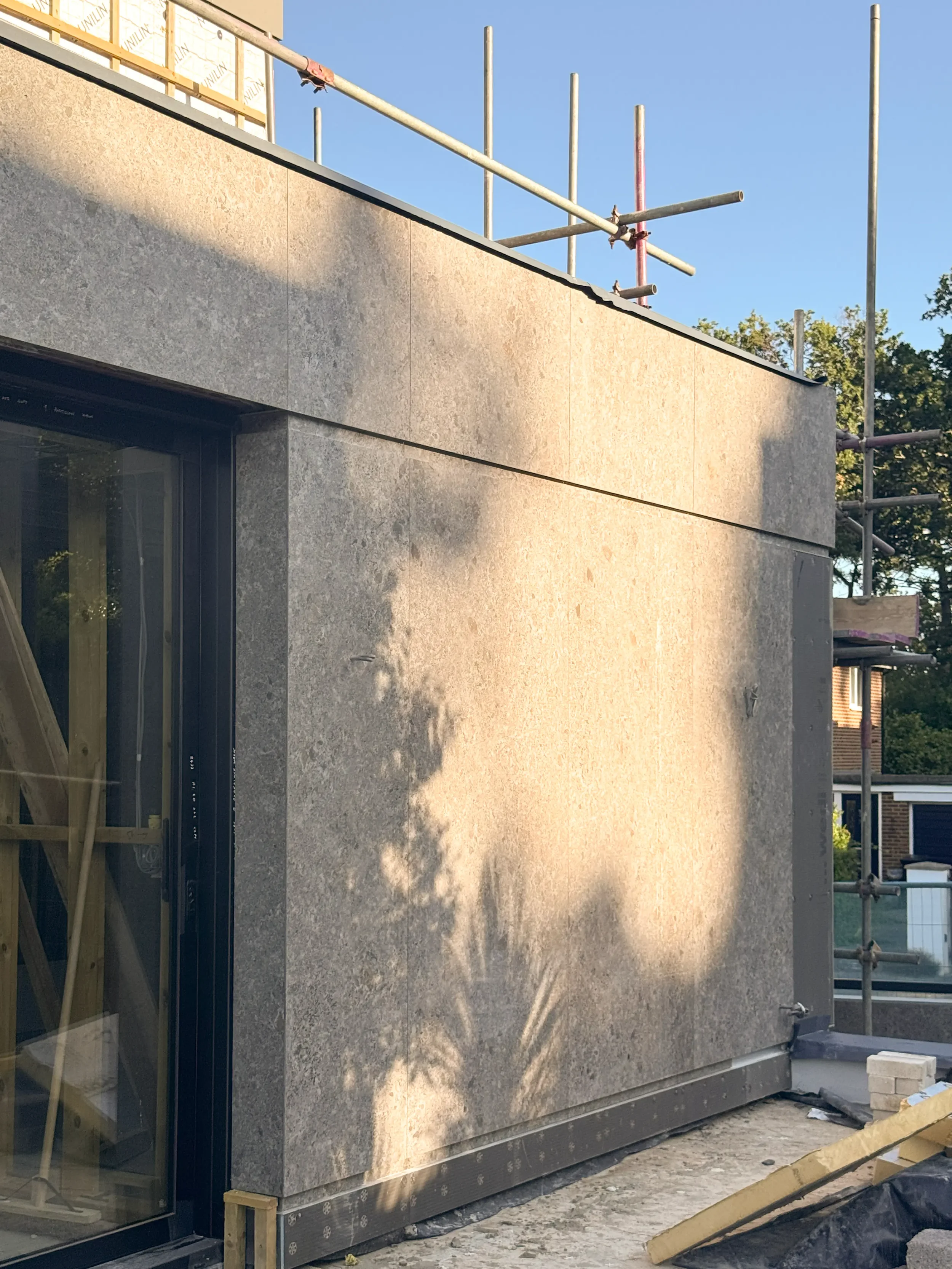 Modern residential build featuring a direct-applied bonded facade system cladded with large-format Ceppo di Gré or stone-effect porcelain slabs, showing exterior construction and scaffolding.