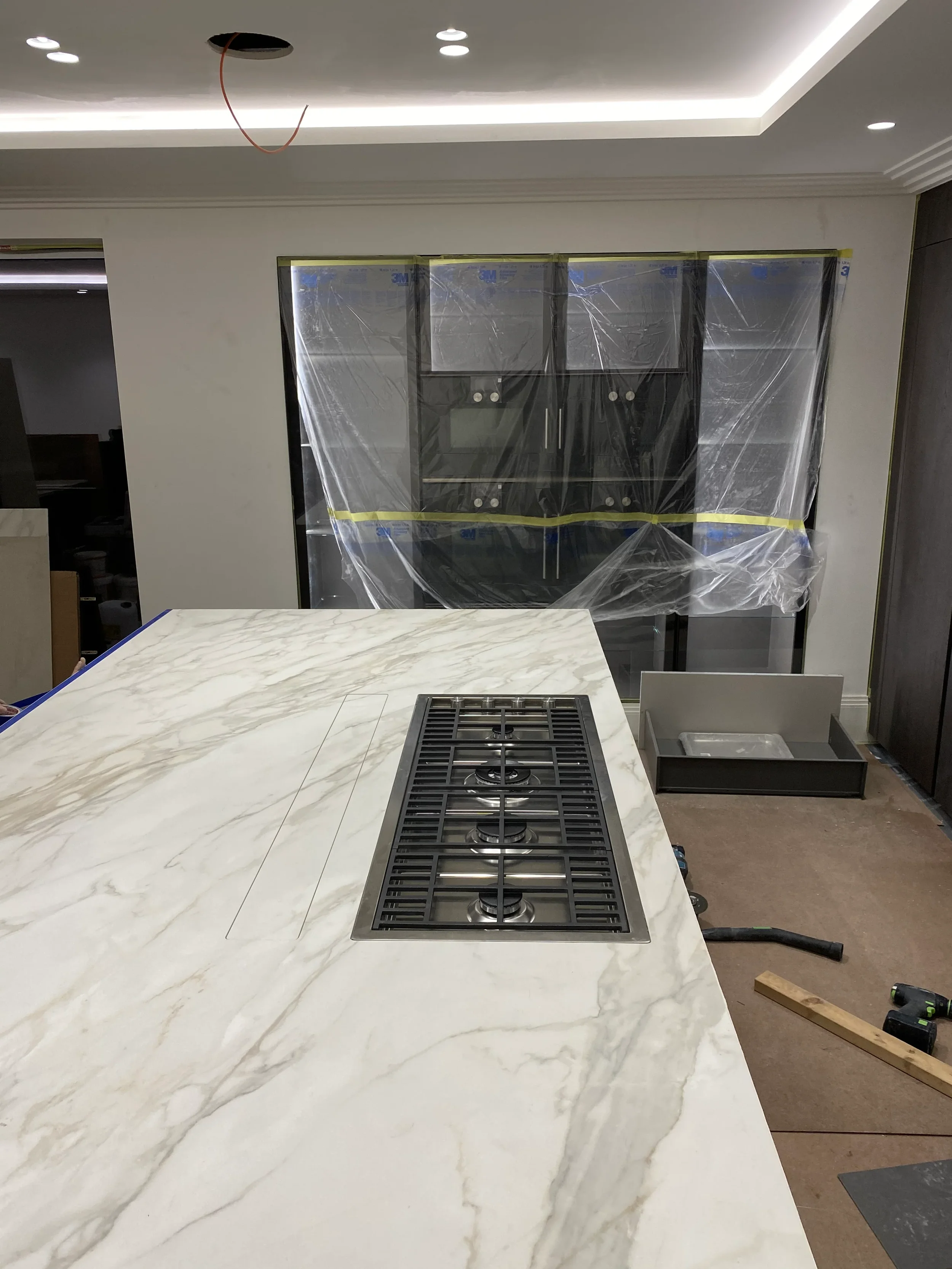 High-end contemporary kitchen under construction, highlighting custom architectural porcelain fabrication with a premium stone-look porcelain worktop, professional linear burner layout, and integrated tall cabinetry covered in protective film.