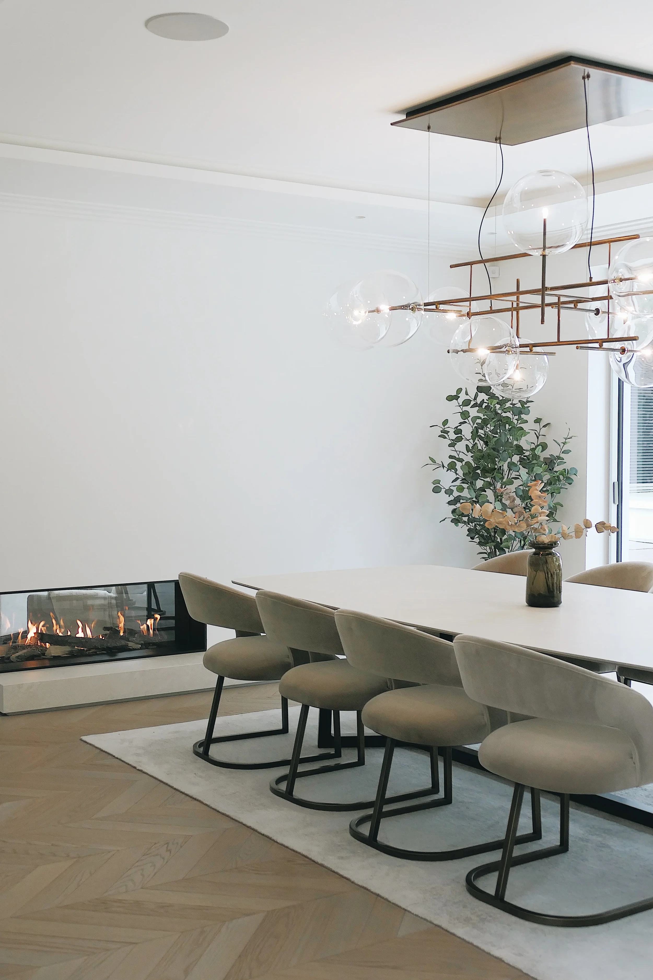 A luxury contemporary dining room featuring a custom-fabricated large format porcelain slab table, velvet dining chairs, a sculptural glass globe chandelier, and a low-profile glass-enclosed linear gas fireplace.