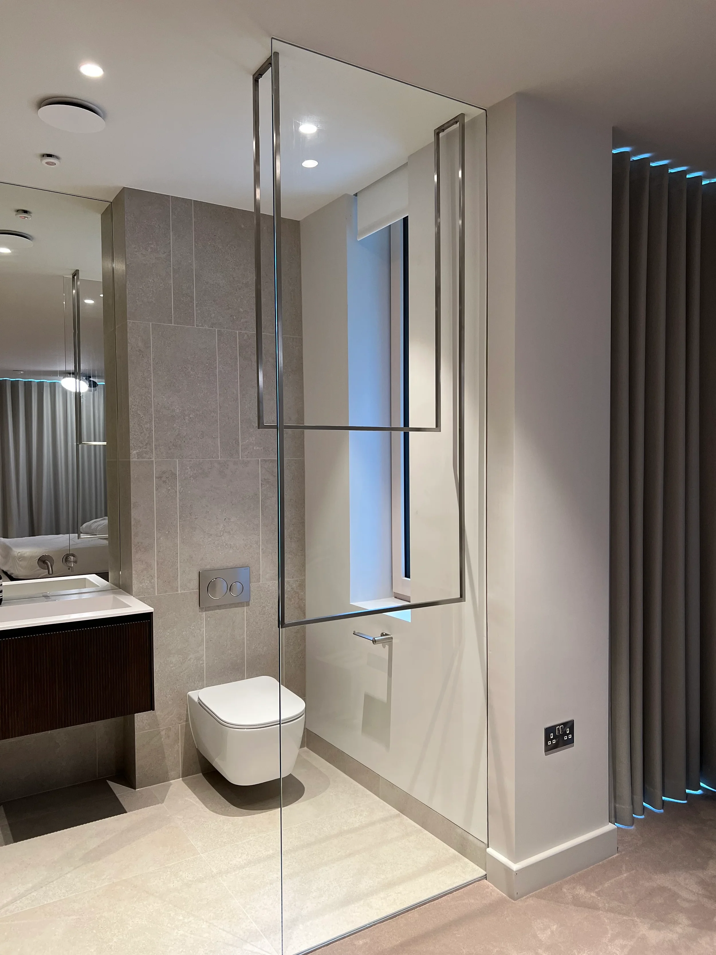 Contemporary bathroom interior with a wall-mounted toilet, large vanity mirror, and a floor-to-ceiling glass partition next to a neutral-toned wall.