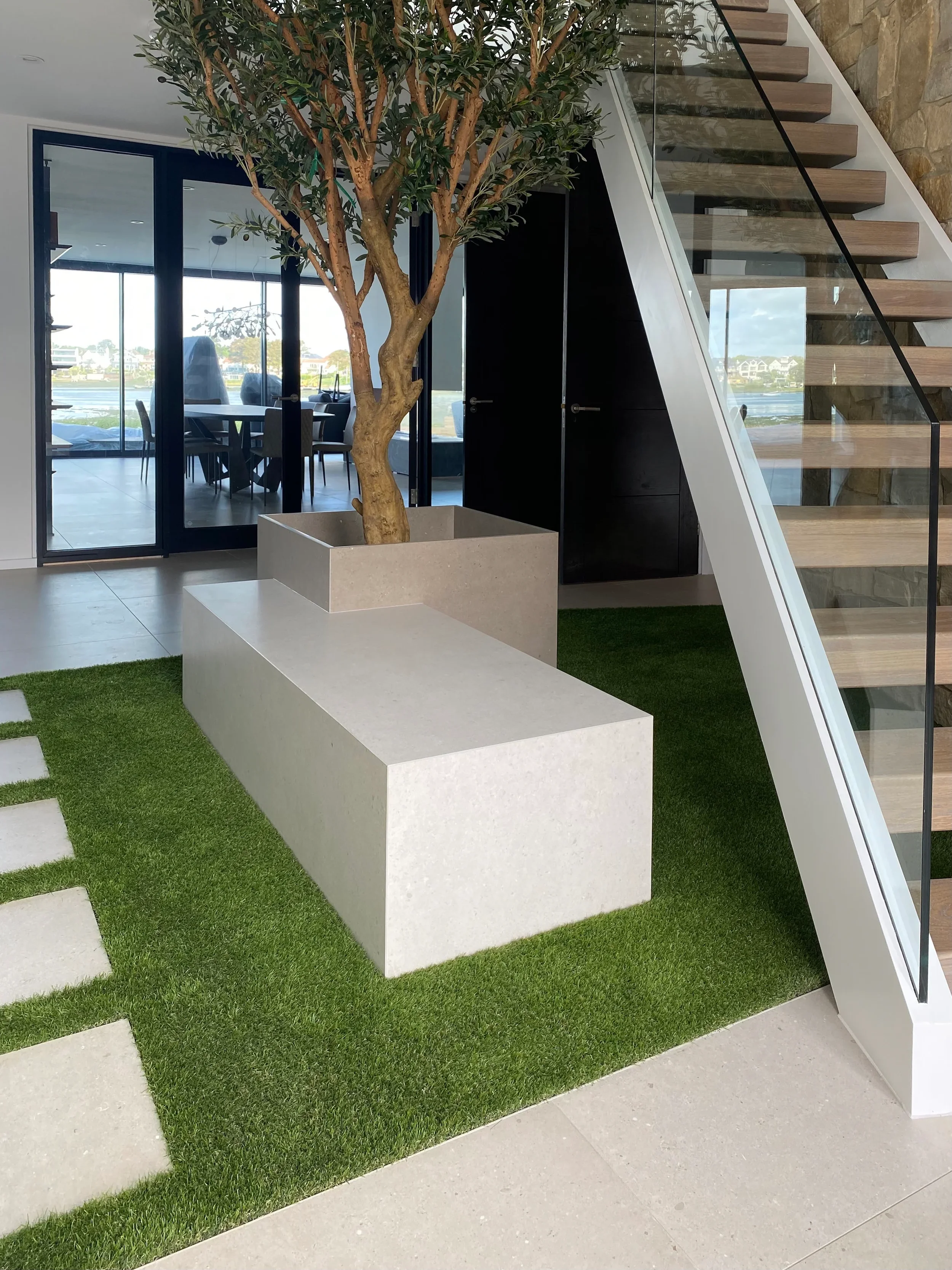 A custom fabricated beige porcelain stone bench and integrated tree planter by PietraCasa set on an indoor green lawn feature in a luxury entrance hallway.