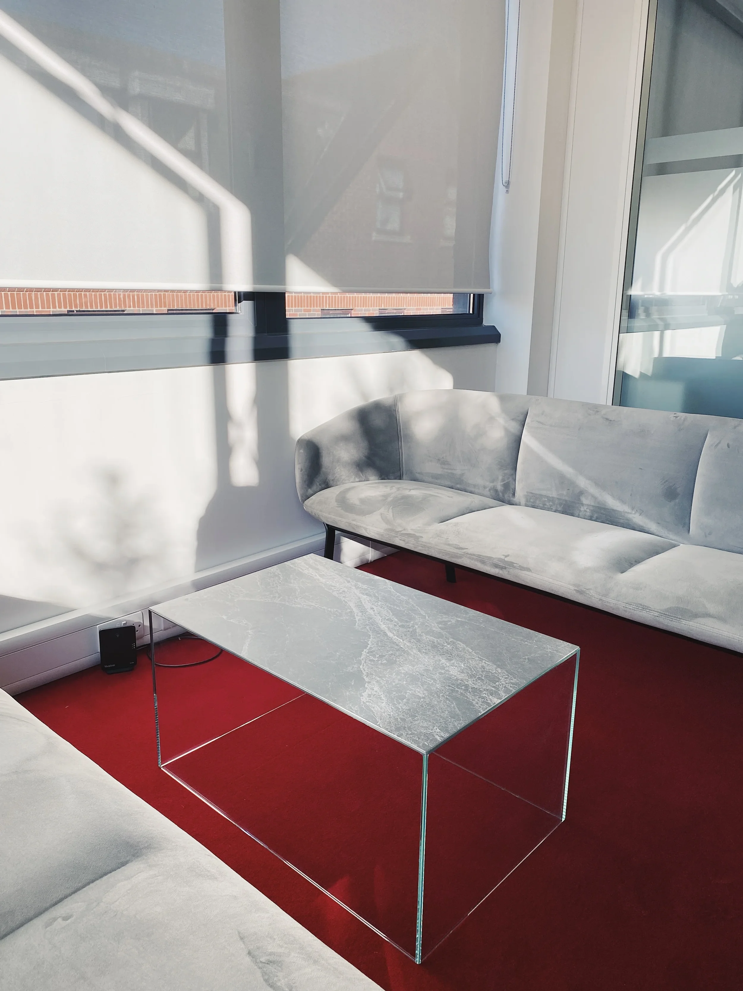 A luxury contemporary coffee table featuring a grey marble-effect porcelain top supported by a minimalist, transparent clear glass base structure.