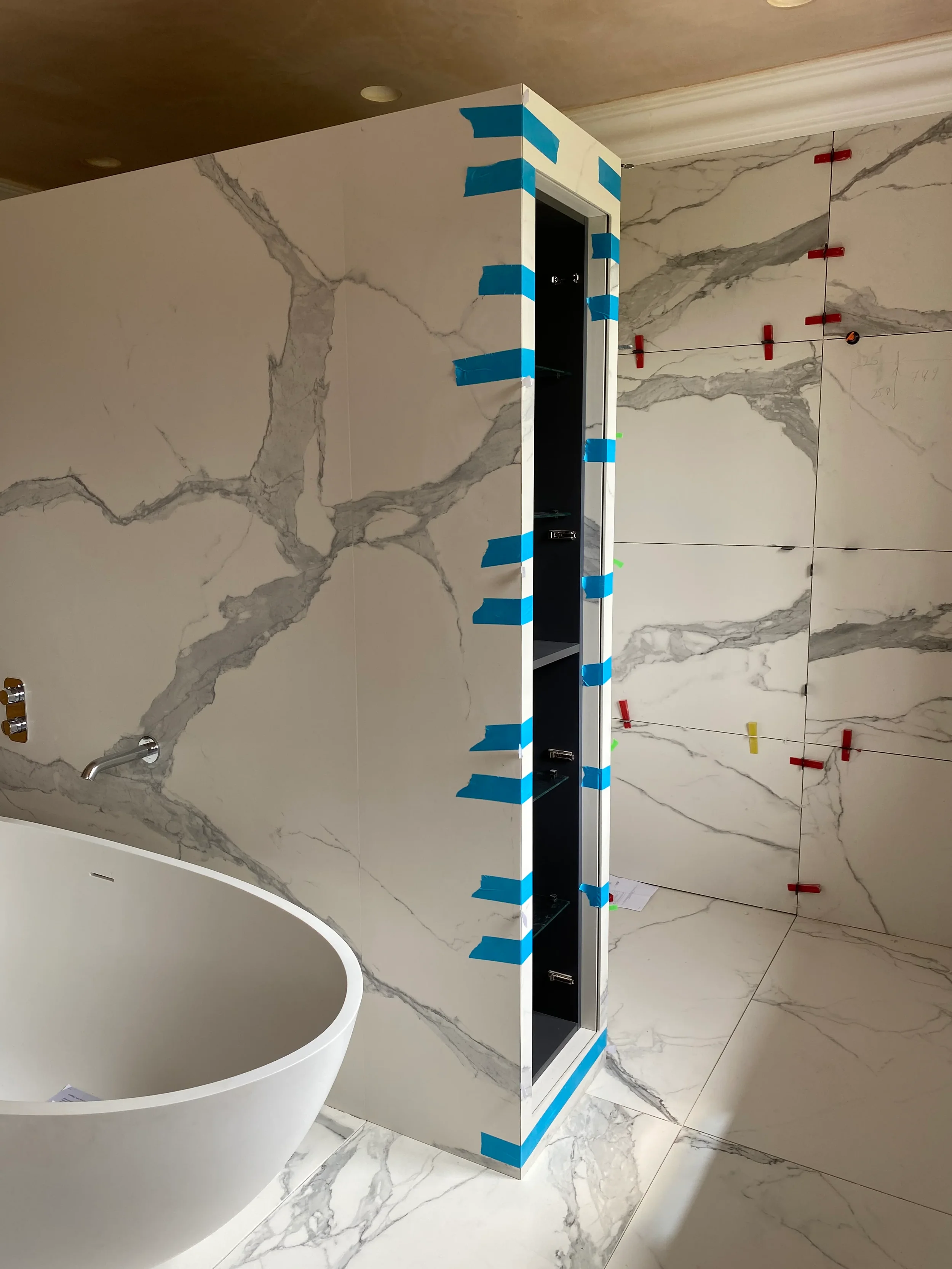 Luxury bathroom under construction featuring bookmatched Calacatta marble-effect porcelain wall and floor tiles by PietraCasa, with leveling spacers and painter's tape on a custom mitred corner.