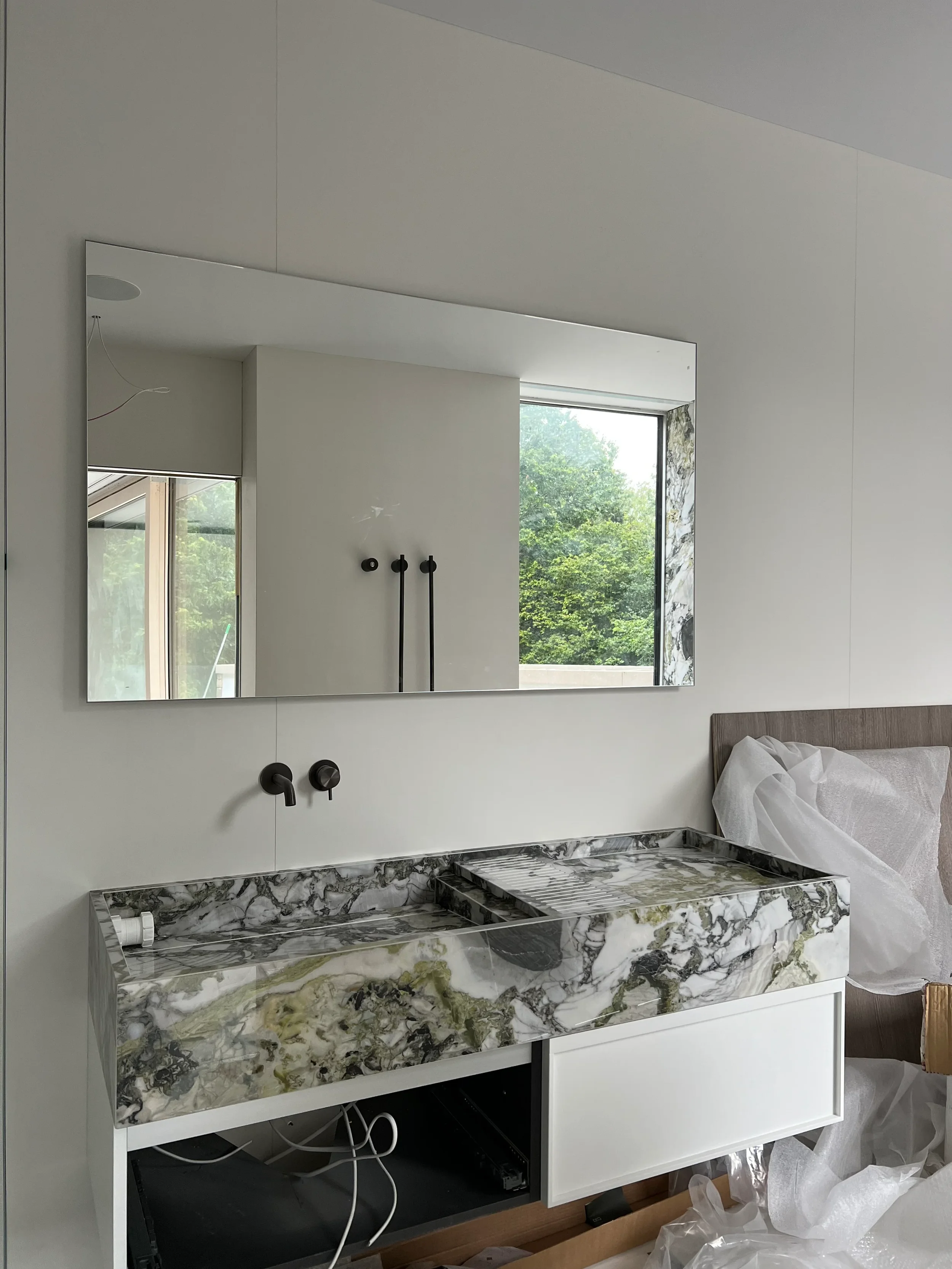 Custom fabricated marble-effect porcelain washbasin with a wall-mounted black mixer tap and a large mirror during installation