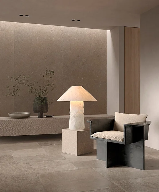 A minimalist interior featuring Kronos Ceramiche porcelain stoneware flooring, an alabaster lamp on a travertine plinth, and a sharp, architectural armchair, available through PietraCasa.