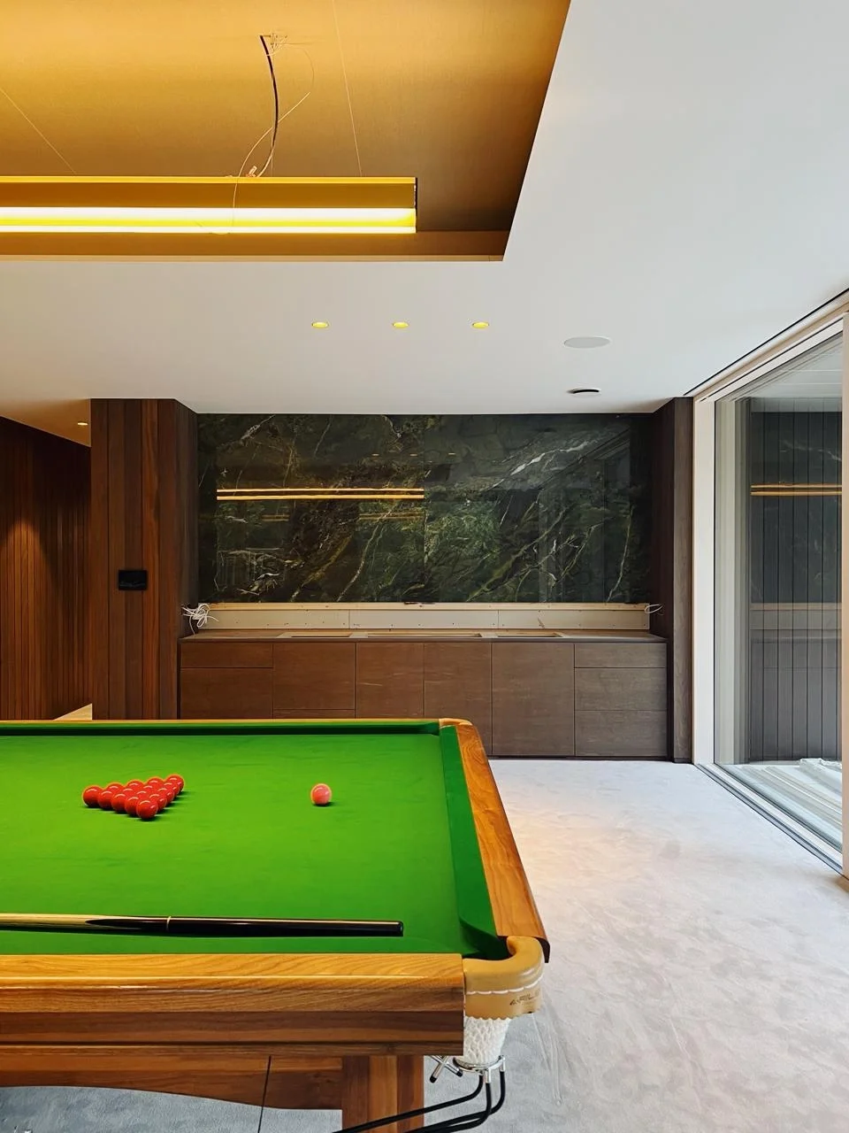 A pool table with red balls and a cue stick in a modern room with wooden and marble walls, large window, and a gold ceiling fixture.