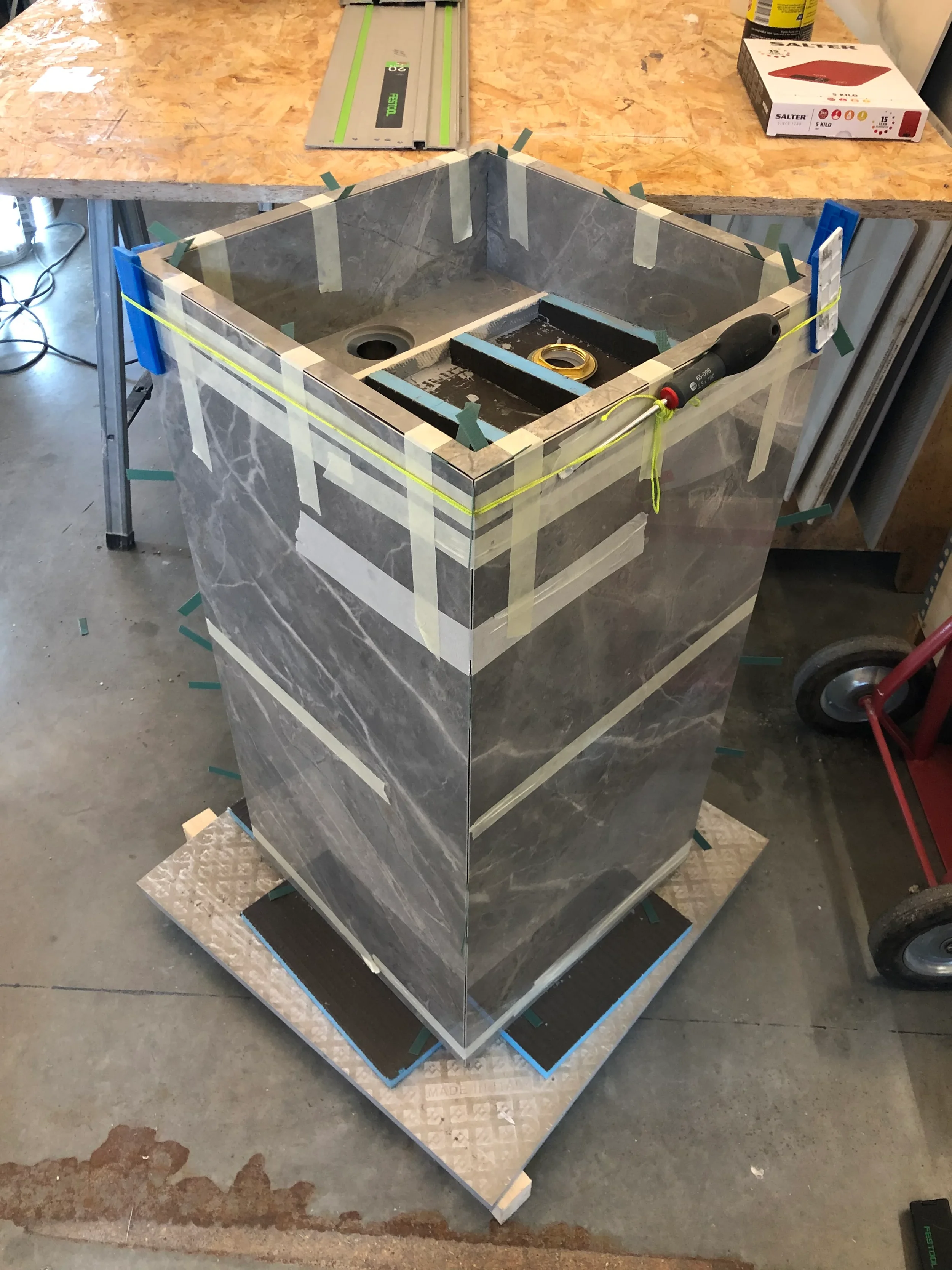 Precision custom architectural porcelain fabrication, highlighting the mitered edge detailing and clamping process of a bespoke grey veined stone-look porcelain pedestal vanity unit during assembly.