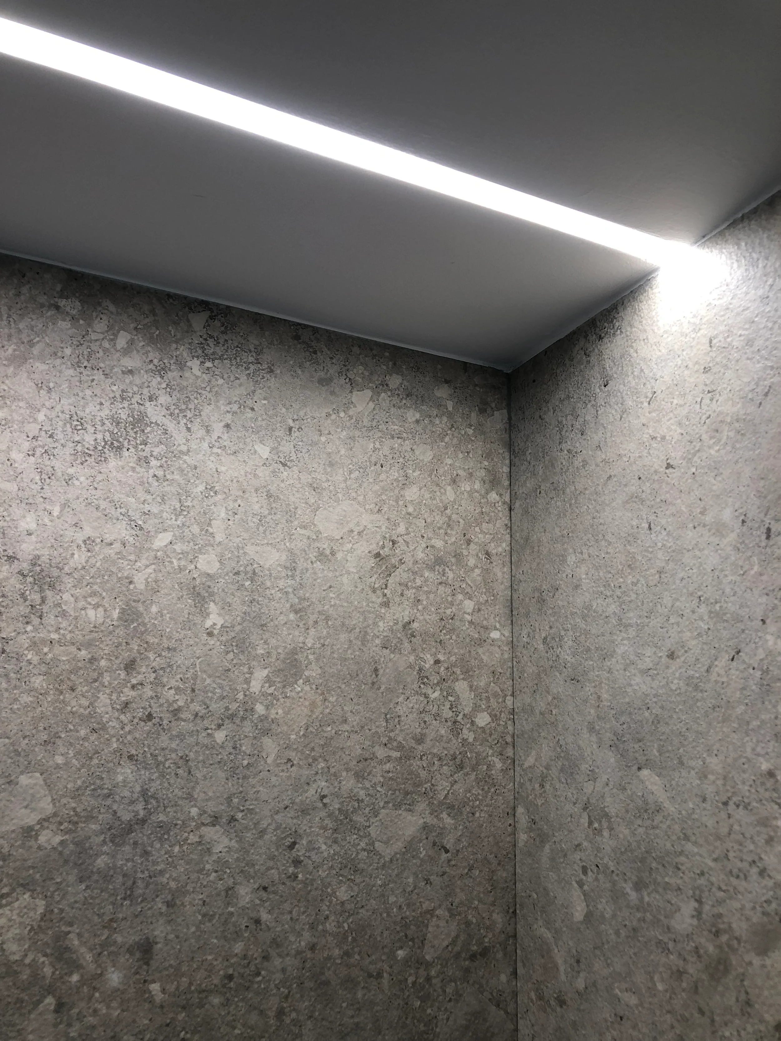 architectural wet room lighting, highlighting a seamless flush-mounted linear LED strip profile illuminating a textured stone-effect large-format porcelain ceiling.