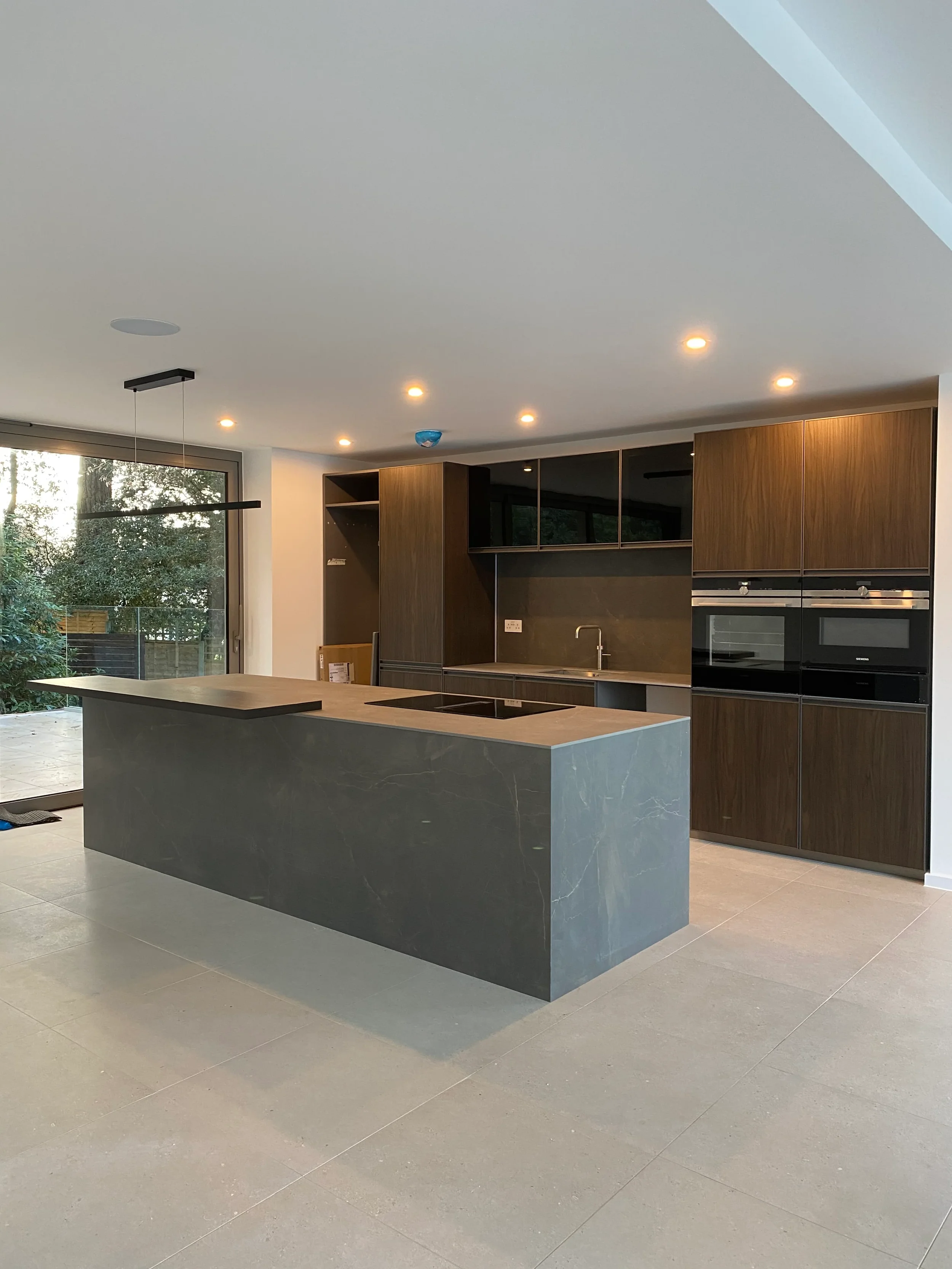 A contemporary luxury kitchen featuring a statement island clad in seamless, large-format dark porcelain, perfectly contrasted with warm wood cabinetry and minimalist design elements.
