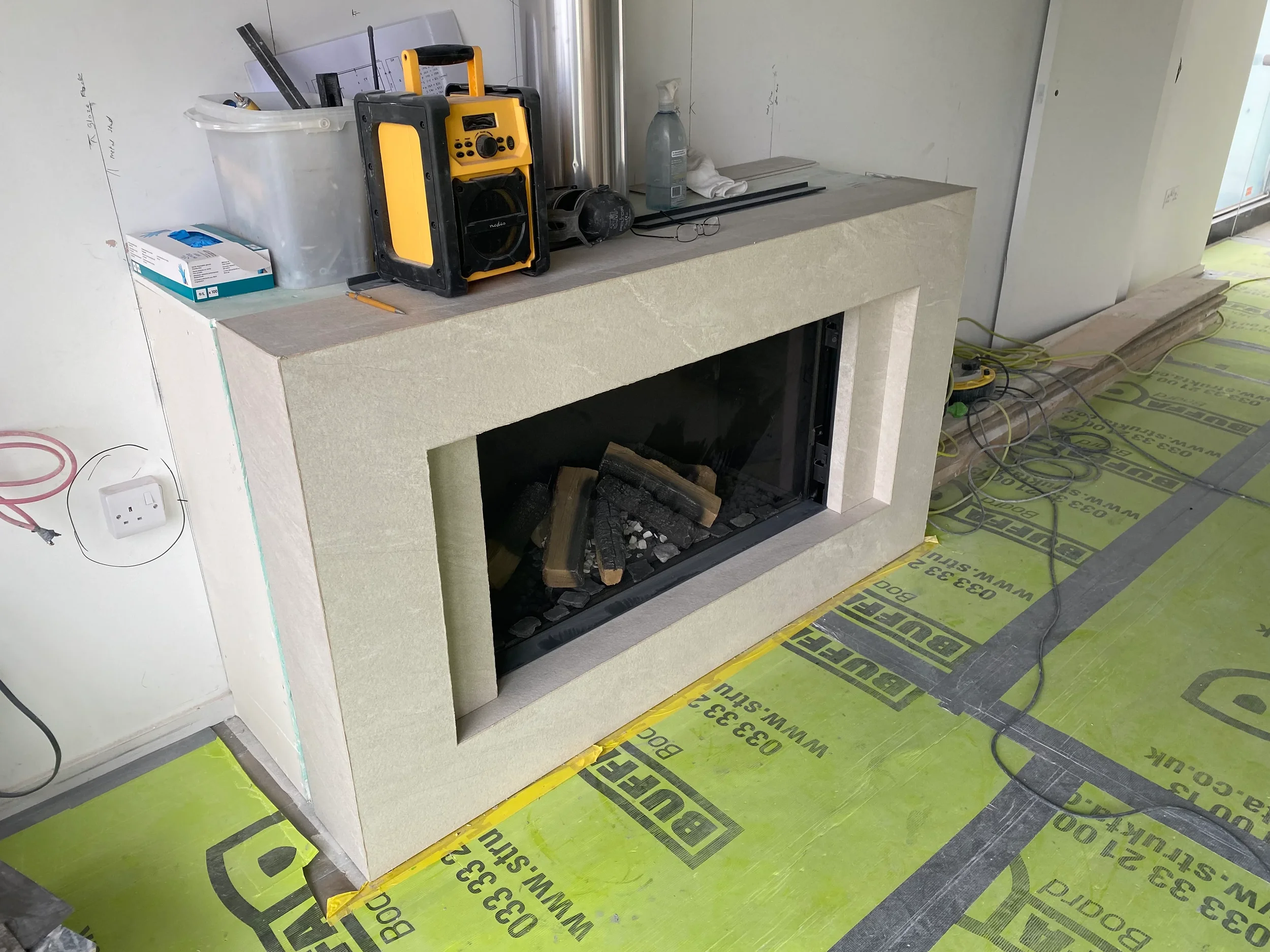 A custom fabricated beige limestone-effect porcelain fireplace surround by PietraCasa, shown mid-installation around a modern gas log burner on a residential construction site.