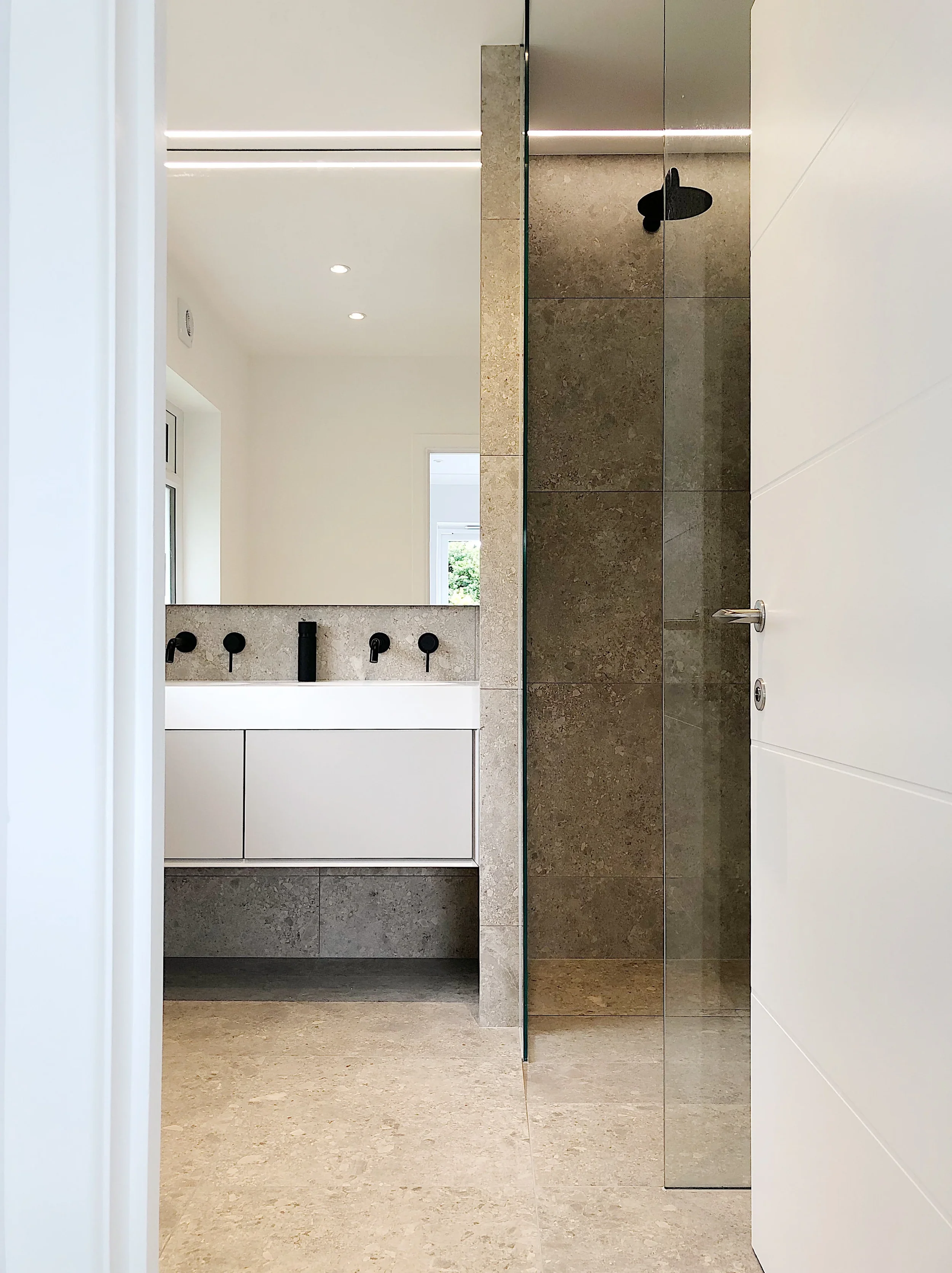 Luxury master en-suite bathroom, featuring a minimalist white floating vanity unit, wall-mounted matt black taps, and a walk-in shower enclosure finished in large-format textured grey Ceppo-style porcelain tiles.