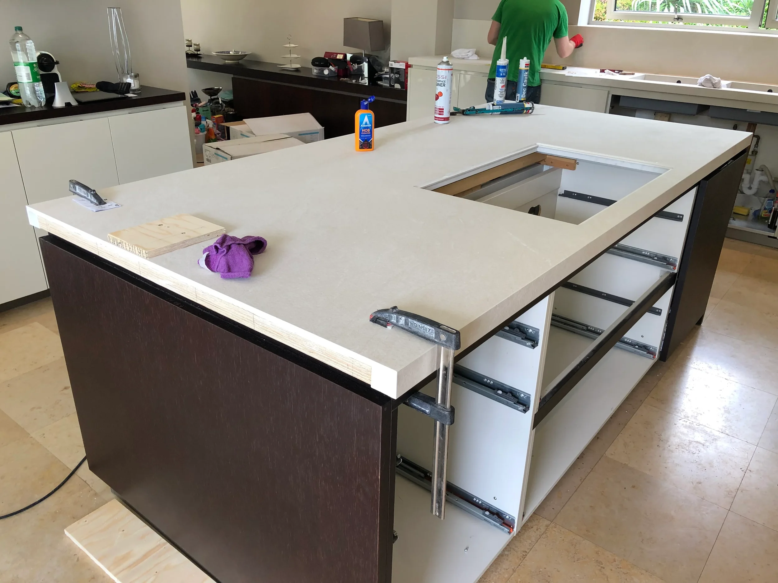 An ongoing luxury kitchen installation featuring a large-format porcelain worktop being fitted onto a custom dark wood kitchen island, showcasing professional craftsmanship and precise detailing.
