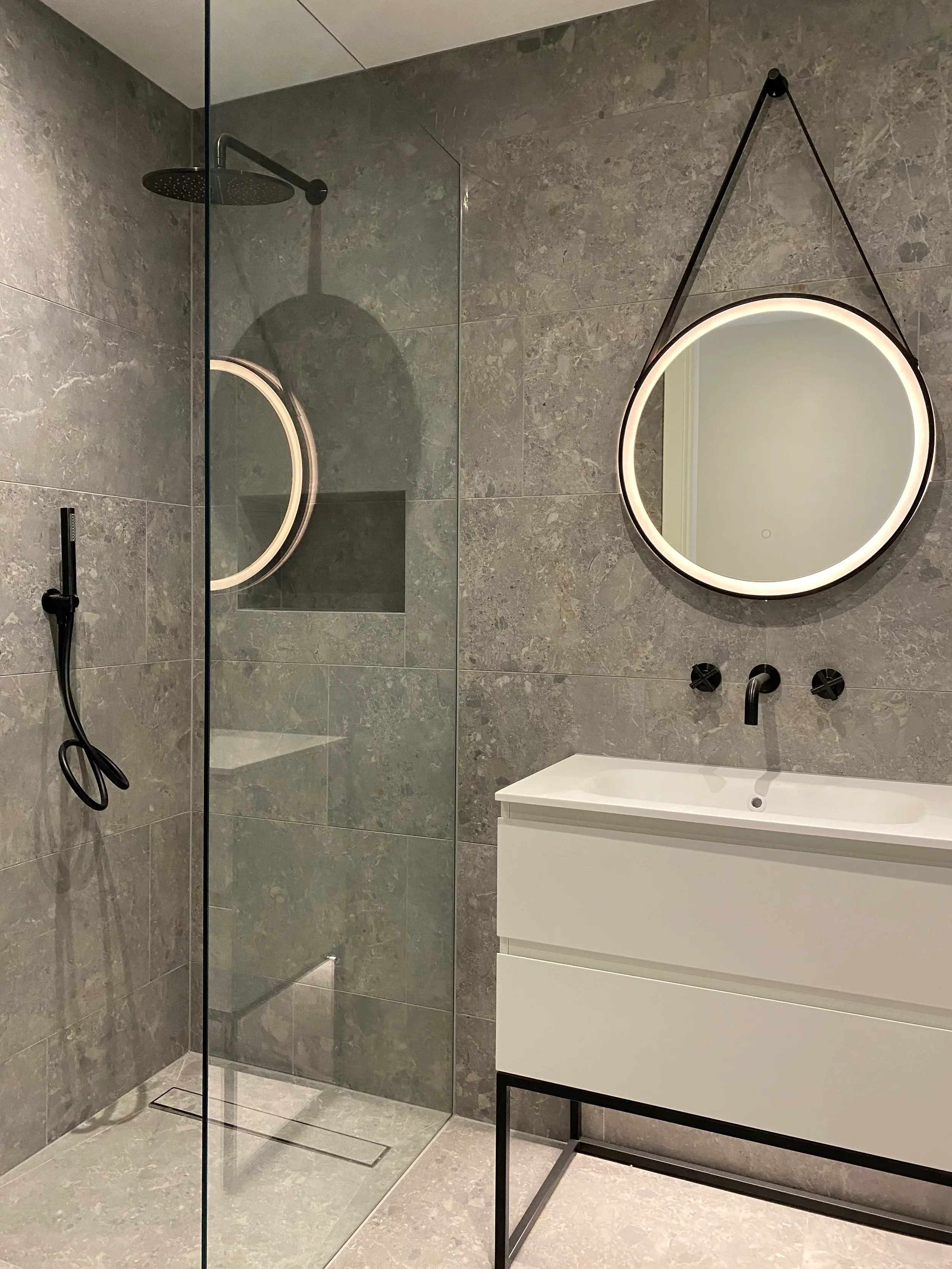 : Contemporary bathroom interior showcasing a walk-in shower, a minimalist white vanity unit, and a circular illuminated mirror against a grey tiled wall.