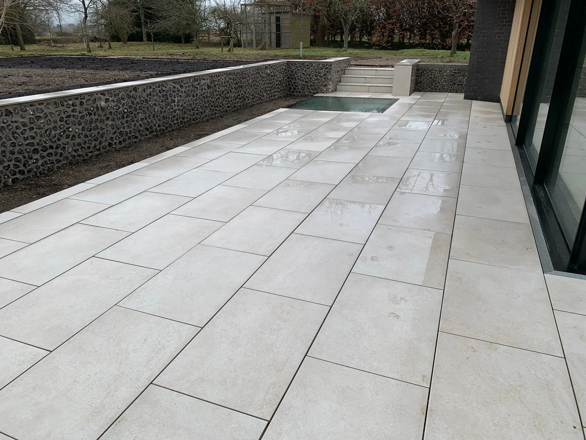 Wide view of an expansive outdoor terrace featuring light-colored, large-format porcelain paving tiles alongside rustic flint masonry walls.