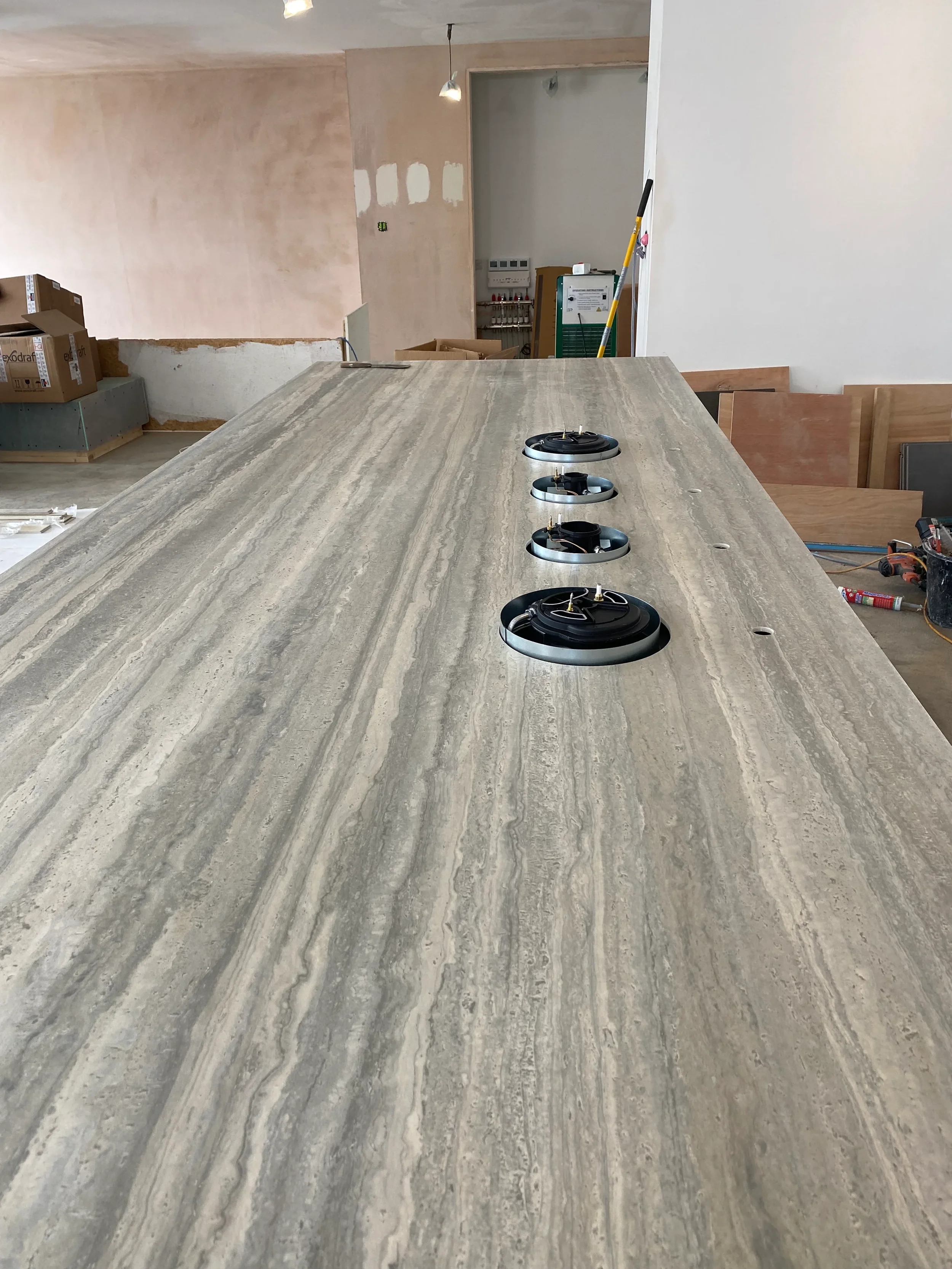 Luxury kitchen island under construction featuring an expansive worktop custom fabricated from vein-cut travertine-effect porcelain slabs, highlighting four precision circular cutouts for an integrated flush-mount gas burner hob system.