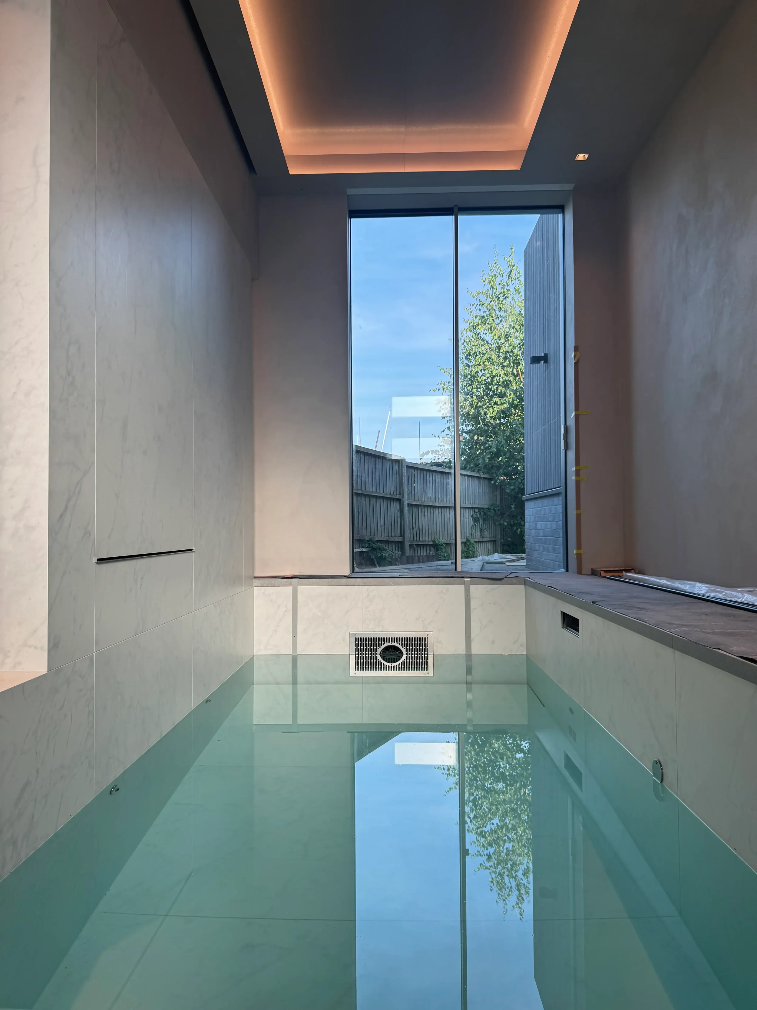 Modern indoor lap pool lined with marble-effect porcelain panels, featuring an architectural coffered ceiling with warm LED lighting.