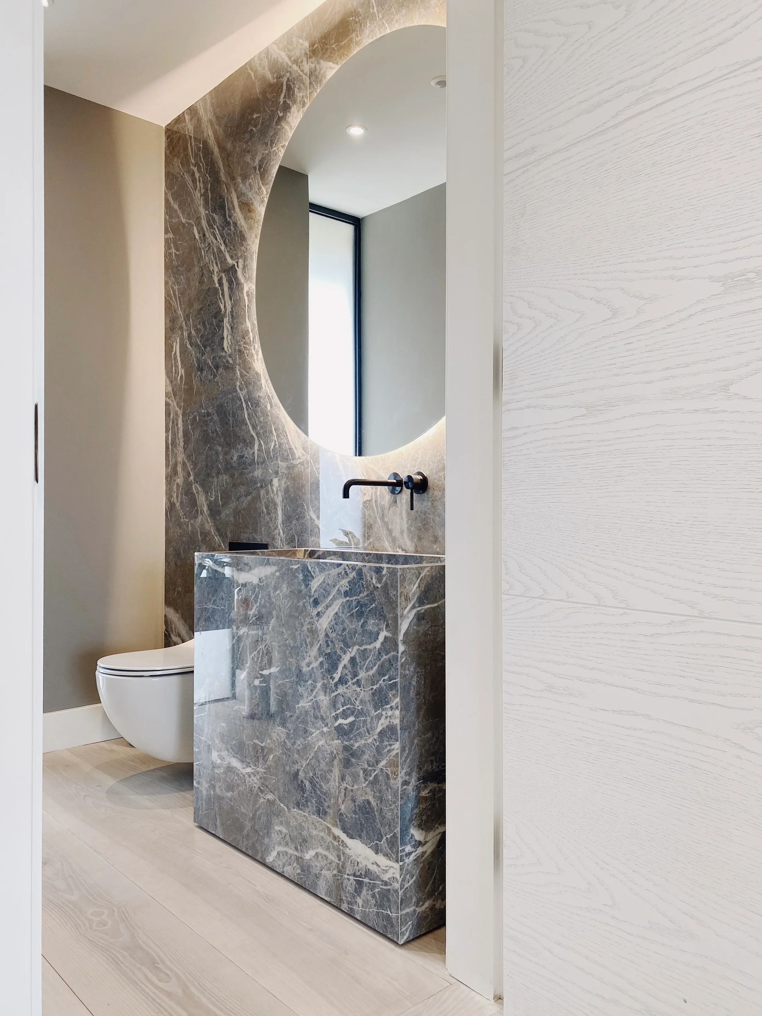 A luxury contemporary cloakroom highlighting a custom-fabricated monolithic vanity basin and matching full-height splashback wall panels in polished grey marble-effect porcelain by PietraCasa.