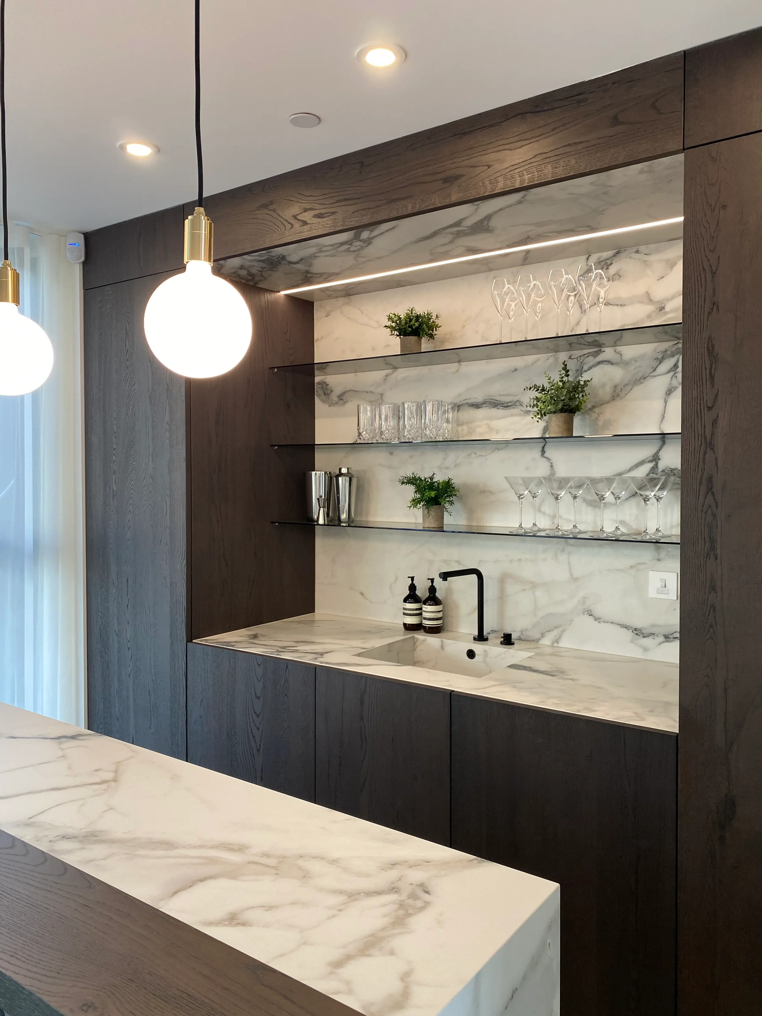 Contemporary home bar and entertaining space featuring a bespoke worktop with a completely integrated 1810 Axix sink and vein-matched splashback custom fabricated from Florim white marble-effect large format porcelain tiles, framed by dark wood cabin