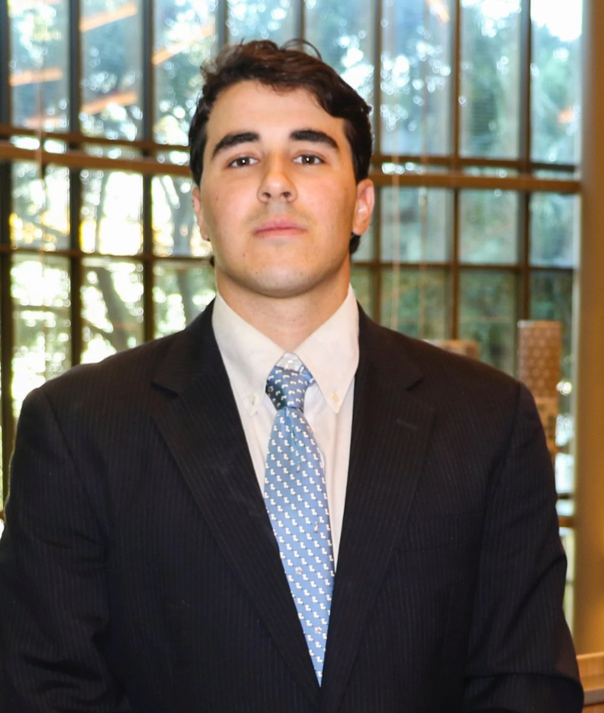 Young man in a dark suit and light-colored shirt with a patterned tie, standing indoors with large windows and a wooded area outside.