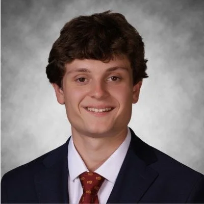A young man with curly brown hair, wearing a dark suit, white shirt, and red tie, smiling against a gray background.