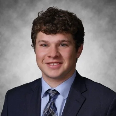 Portrait of a young man with curly hair wearing a navy suit, light blue shirt, and plaid tie, smiling against a gray background.