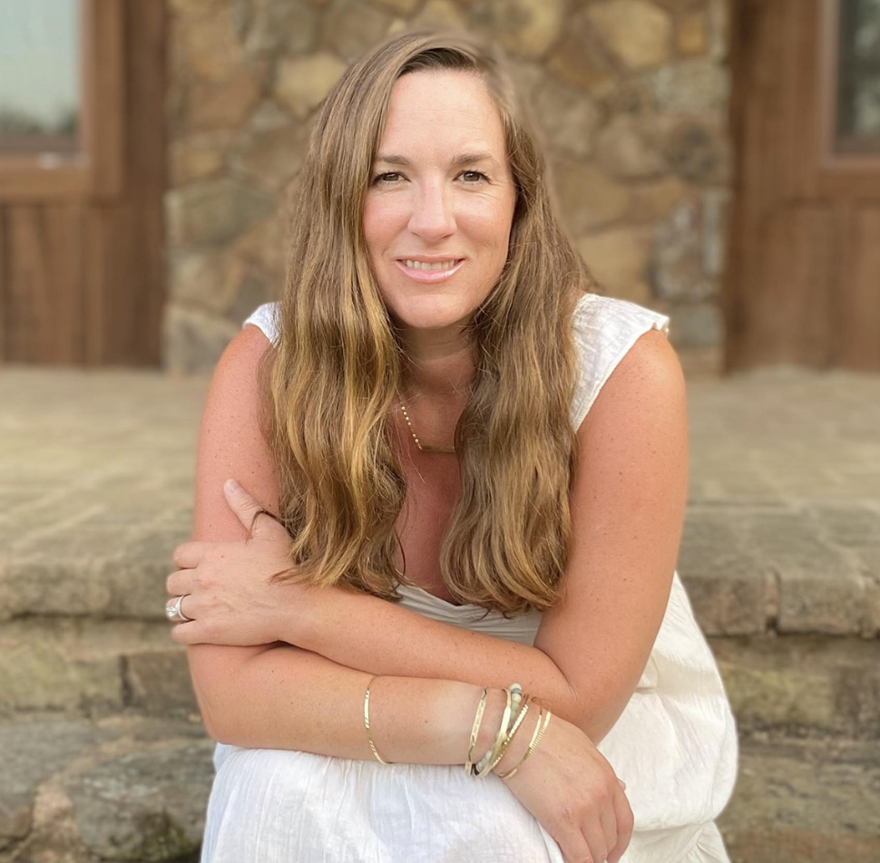 A woman with long wavy blonde hair, wearing a white sleeveless dress, is sitting outdoors in front of a stone wall. She is smiling at the camera with her arms crossed in her lap, accessorized with bracelets, rings, and a delicate necklace.