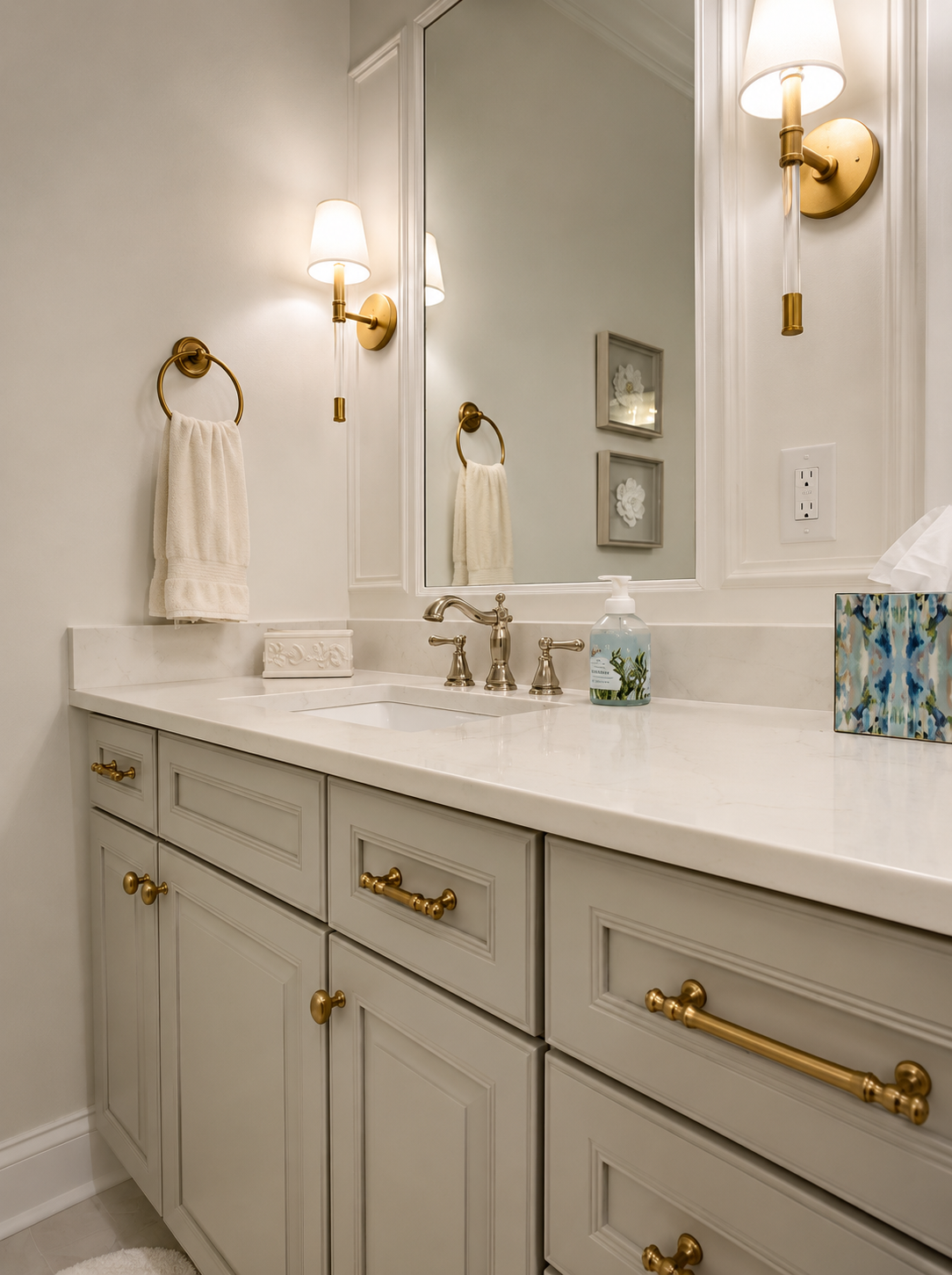 Bathroom vanity with white cabinetry, gold hardware, and a white face marble countertop. There are two wall sconces with white shades, a large mirror, and framed floral artwork on the wall. Items on the counter include a soap dispenser, tissue box, and a small decorative box.