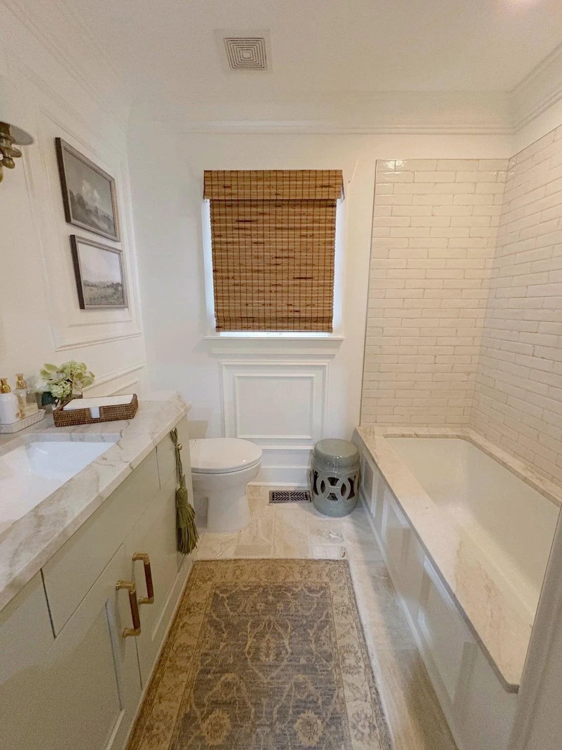 A bathroom with a bathtub, toilet, vanity with sink, and a window with a bamboo shade. The room has white walls, a beige rug, and decorative touches like framed art and a small stool.