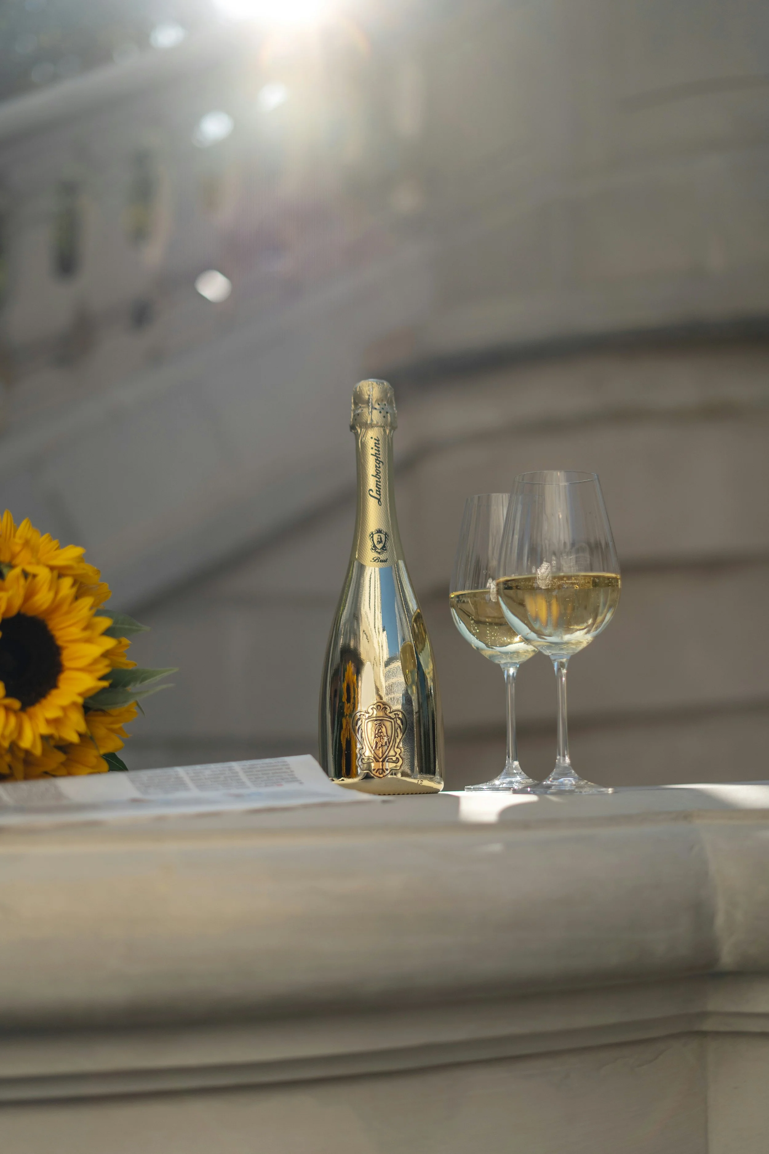 A bottle of Lamborghini champagne and two glasses of white wine on a white surface next to a sunflower on the left, with a blurred background and sunlight shining from above.