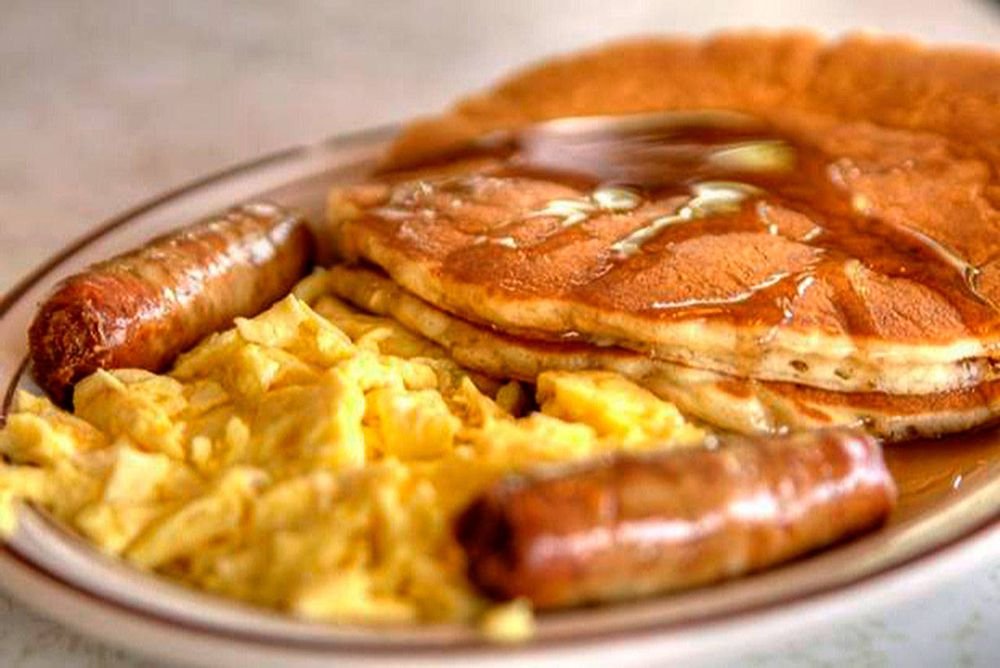 A breakfast plate with scrambled eggs, two sausages, and two pancakes drizzled with syrup.