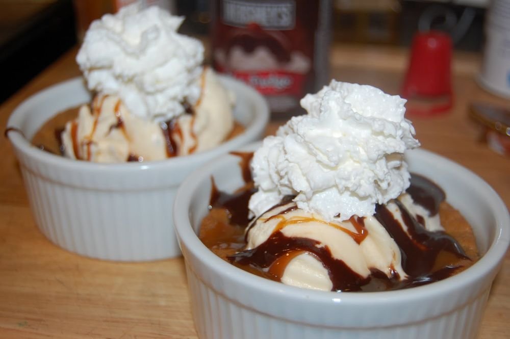 Two bowls of vanilla ice cream topped with whipped cream and drizzled with chocolate and caramel sauce, served on a wooden table.