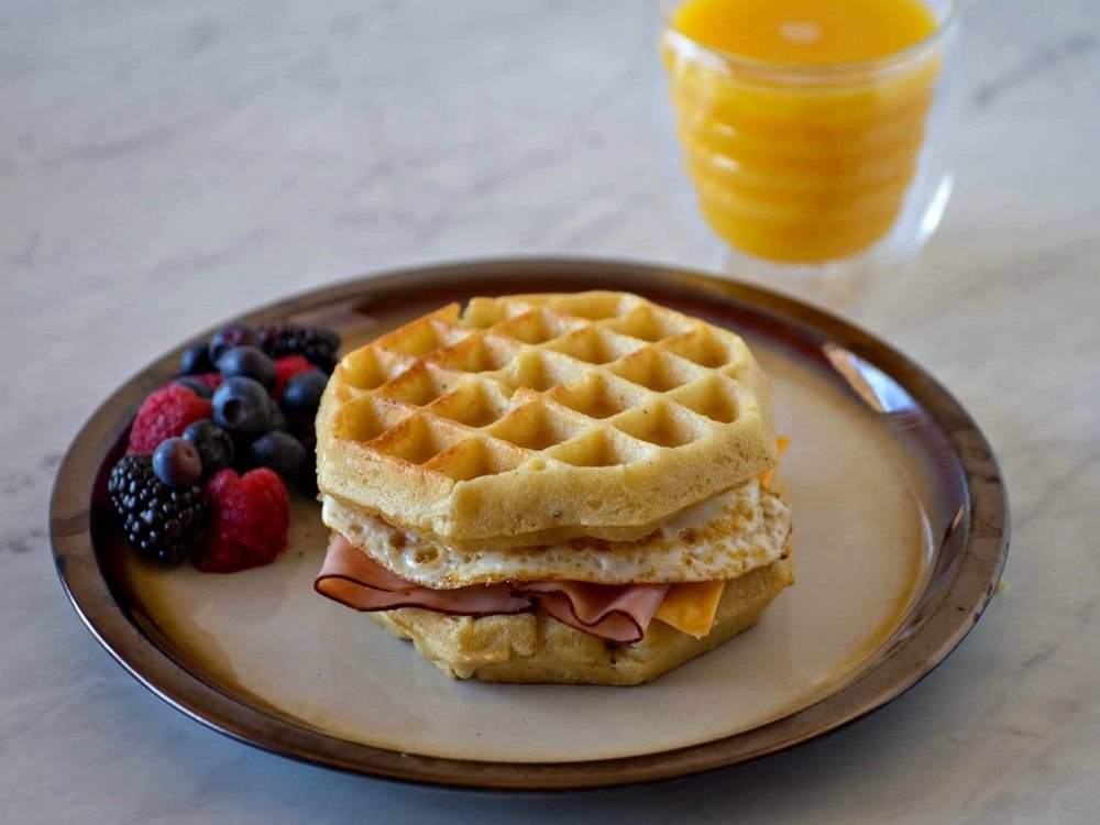 Waffle sandwich with ham, cheese, and egg served with mixed berries and a glass of orange juice.