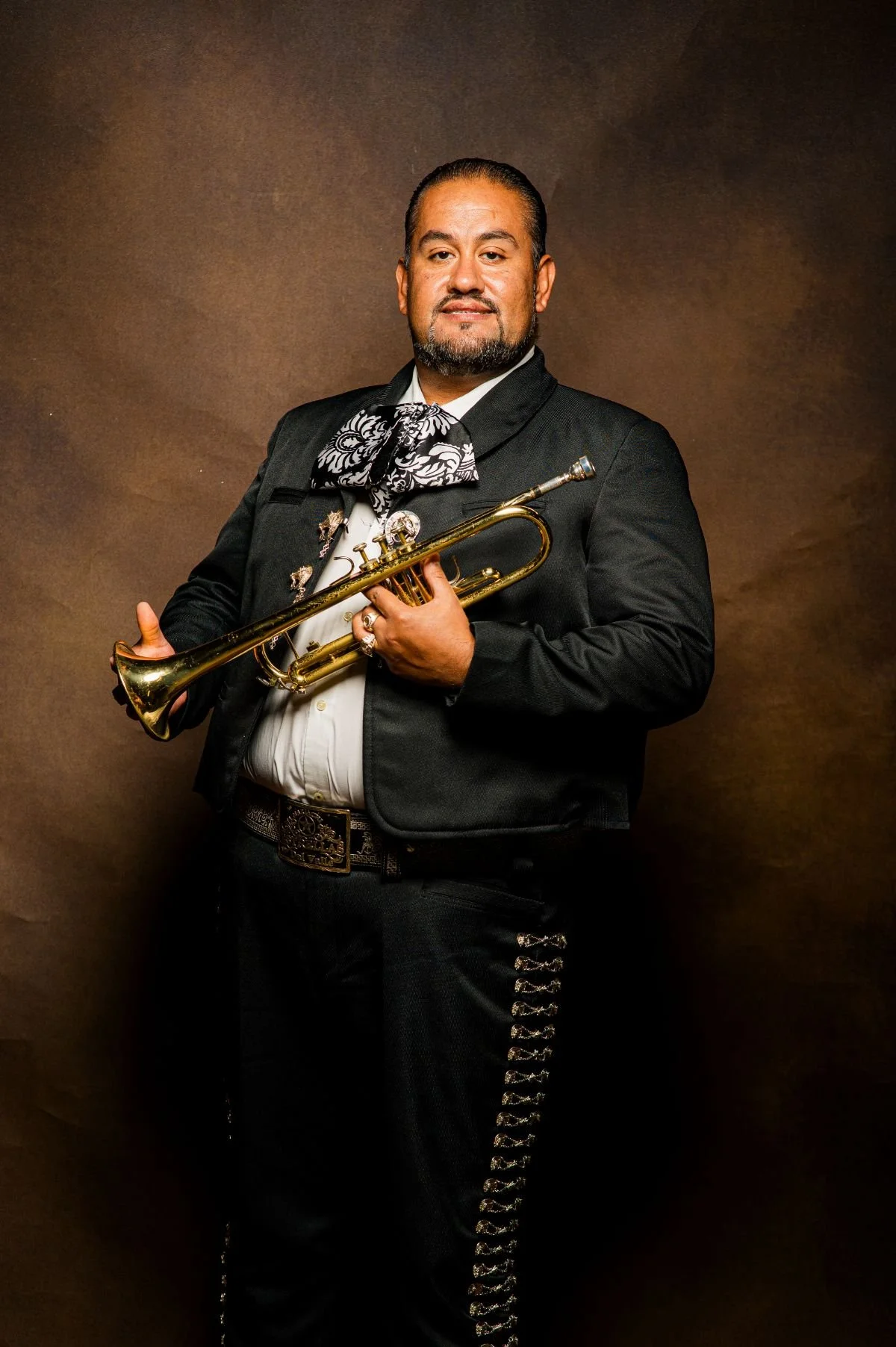A man wearing a traditional Mexican Mariachi outfit holding a trumpet, standing against a brown background.